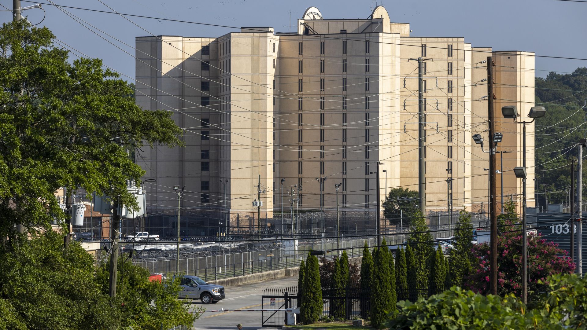 Large beige prison building with tall walls, barbed-wire fences, power lines, and a gated entrance. Green trees and shrubs surround the area, with a person walking in front.