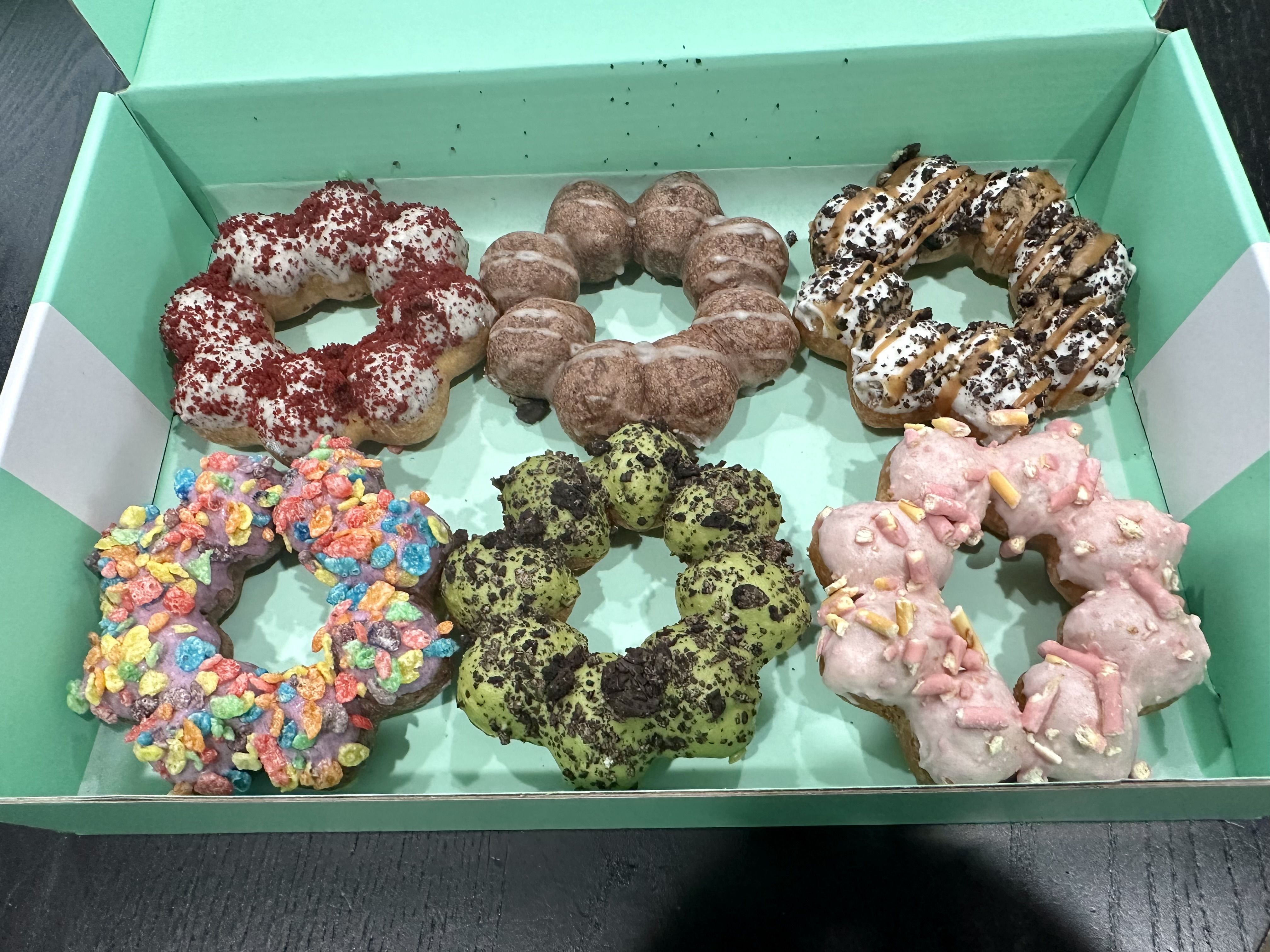 Mochi-style doughnuts, six of them, in colors ranging from green to pink to red and white, with one covered in colorful cereal.