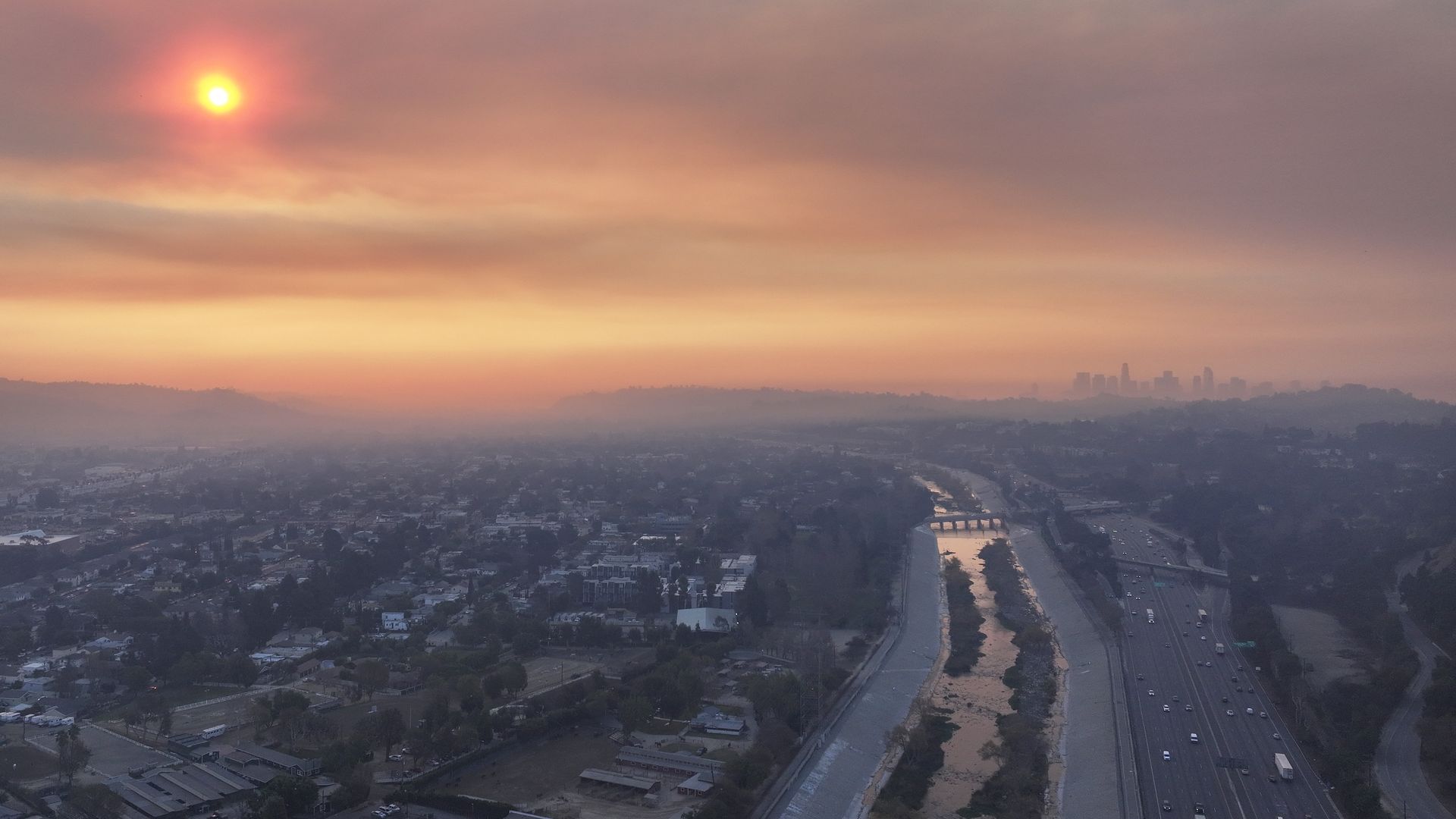 A view of a smoky sky over a freeway through Los Angeles with the sun shining bright reddish orange because of the smoke.