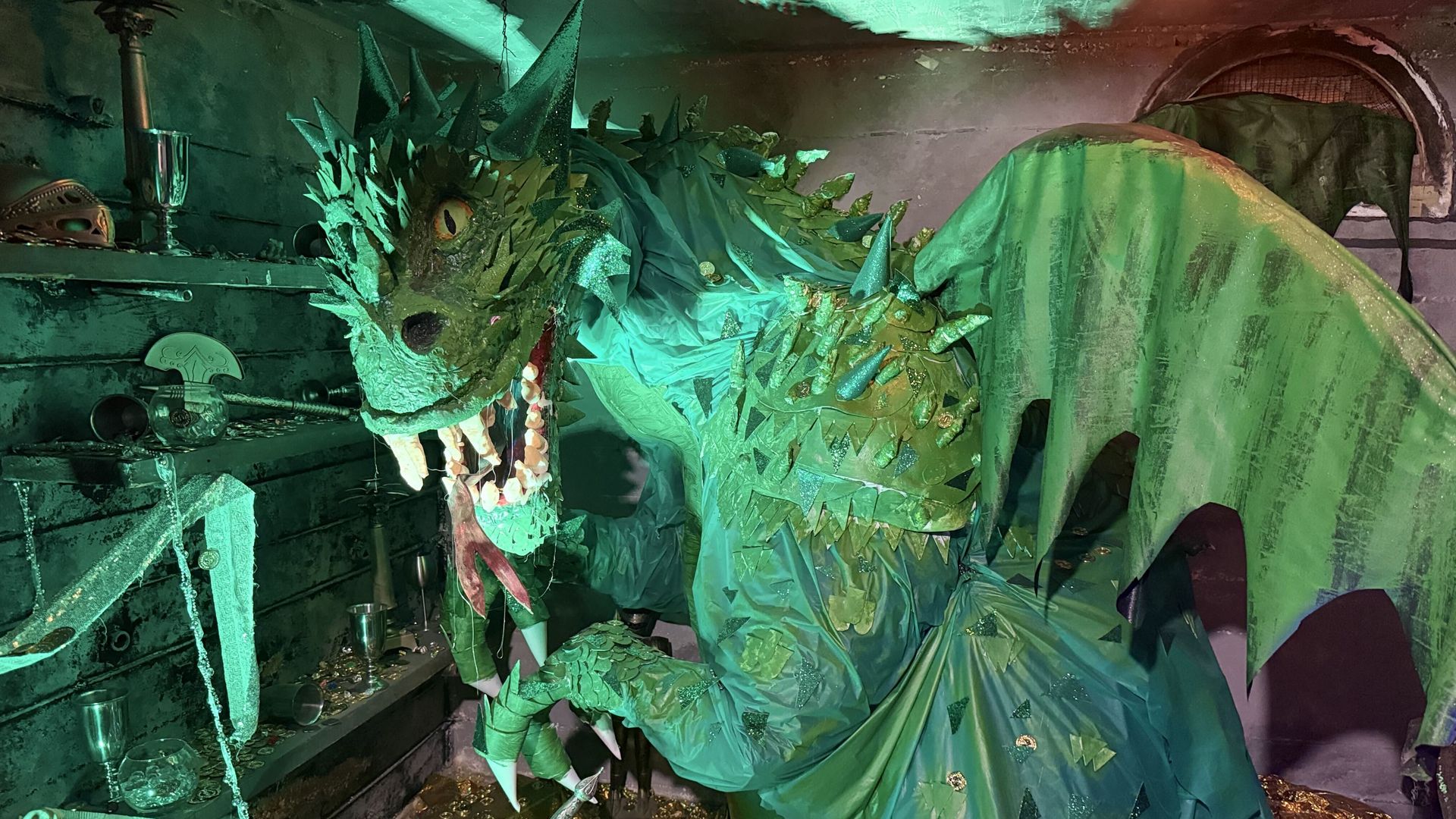 Green-lit treasure room with a large dragon sculpture, spiky emerald scales, and an open mouth full of teeth. Surrounding are scattered gold coins, goblets, a helmet, and dim shelves.
