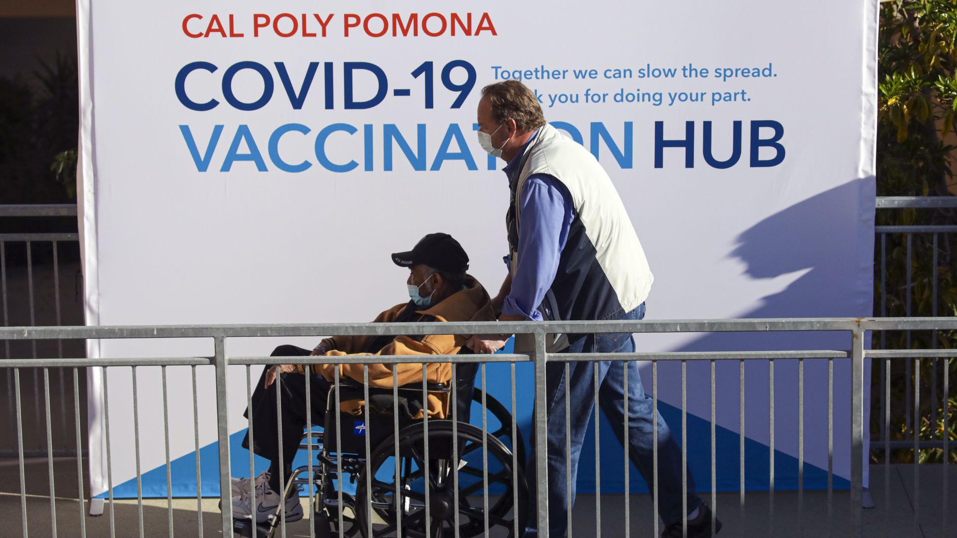 A man is pushed in a wheelchair in front of a sign that reads "Covid-19 vaccination hub"