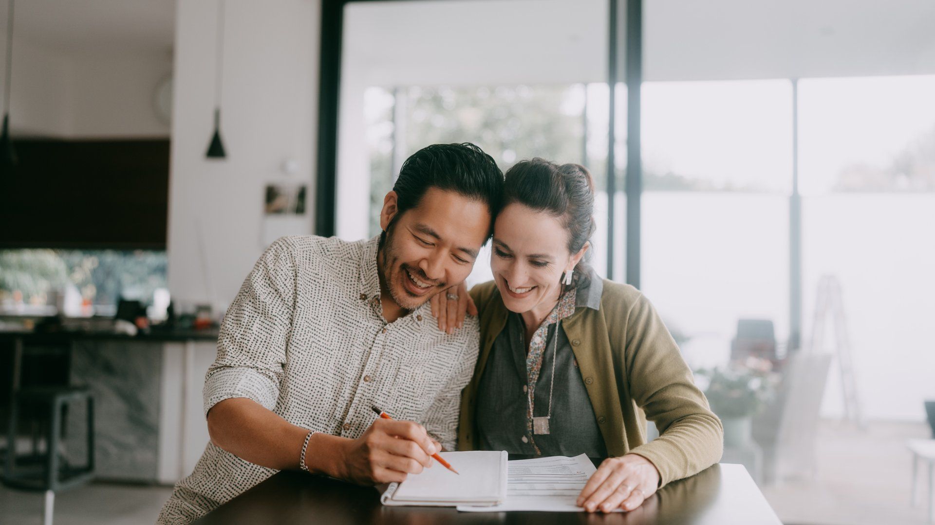 Smiling couple sitting at a table in a bright room, writing on a notebook with documents in front of them, showing engagement and collaboration.
