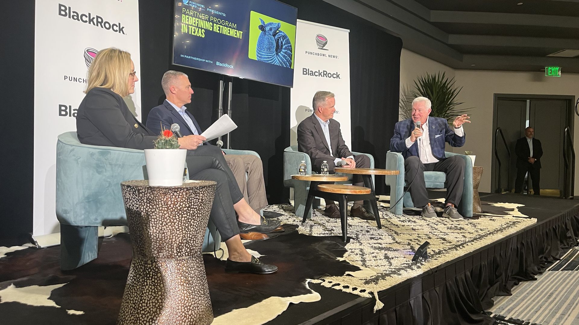 Panel of four people seated on a stage discussing "Redefining Retirement in Texas," with Punchbowl News and BlackRock banners, a screen showing program details, and a small cactus on a side table.