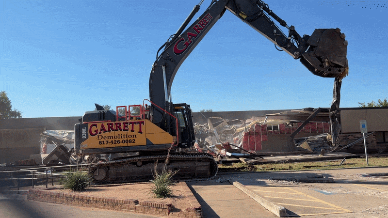Large yellow and black excavator labeled sits amid rubble and a partially demolished building.