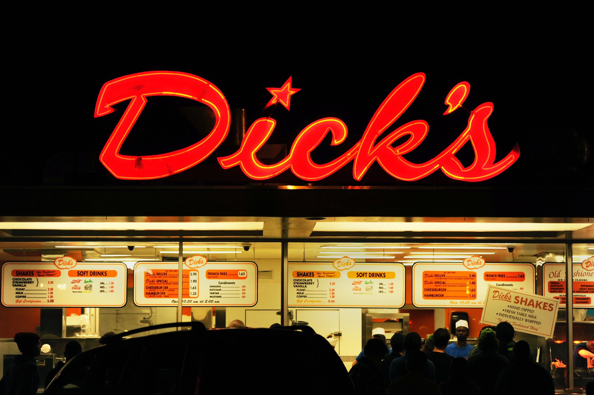 A neon sign that says "Dick's" in cursive red letters, with a star on the 'I," above a drive-in restaurant with menu boards and people lined up to order.