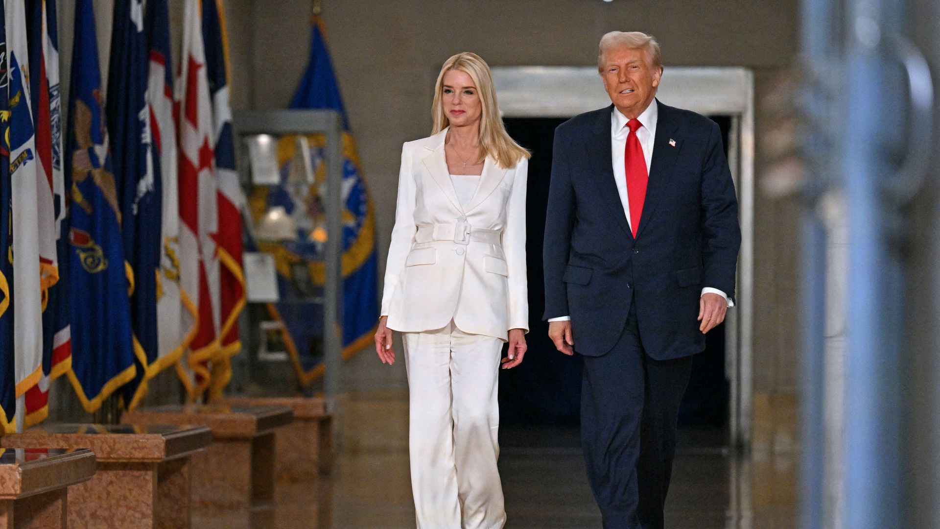 Donald Trump — wearing a red tie, a dark suit and a white collared shirt — walks alongside Pam Bondi, who is wearing a white pantsuit.