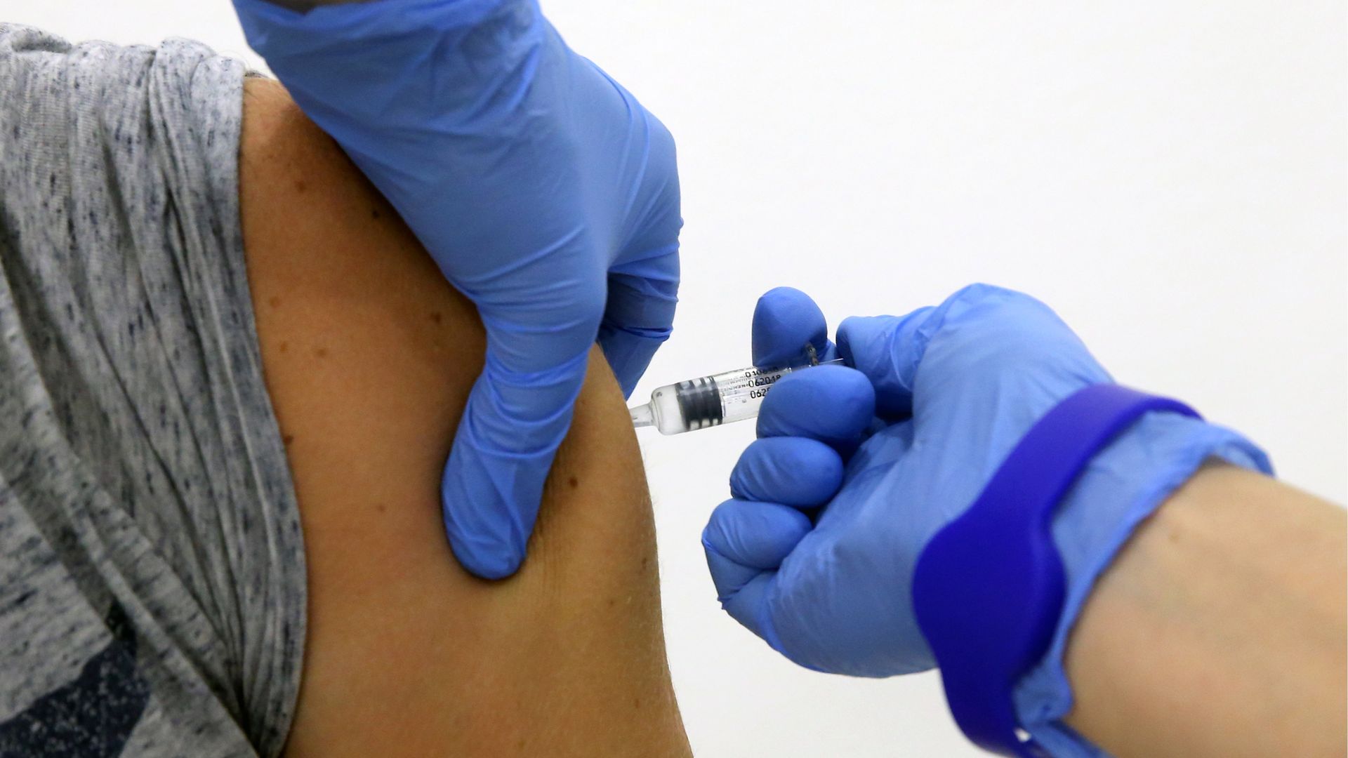 In this image, two gloved hands administer a shot into someone's shoulder.