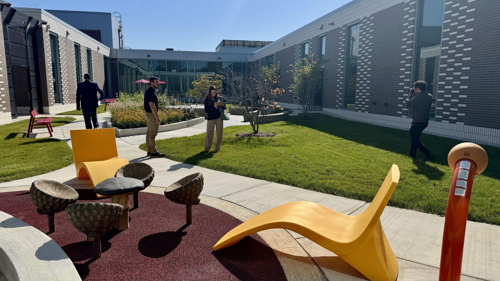 Sunlit courtyard between brick buildings. A man in black, a woman reading, and a photographer on the right. Bright yellow lounge chairs and round woven stools dot the lawn.