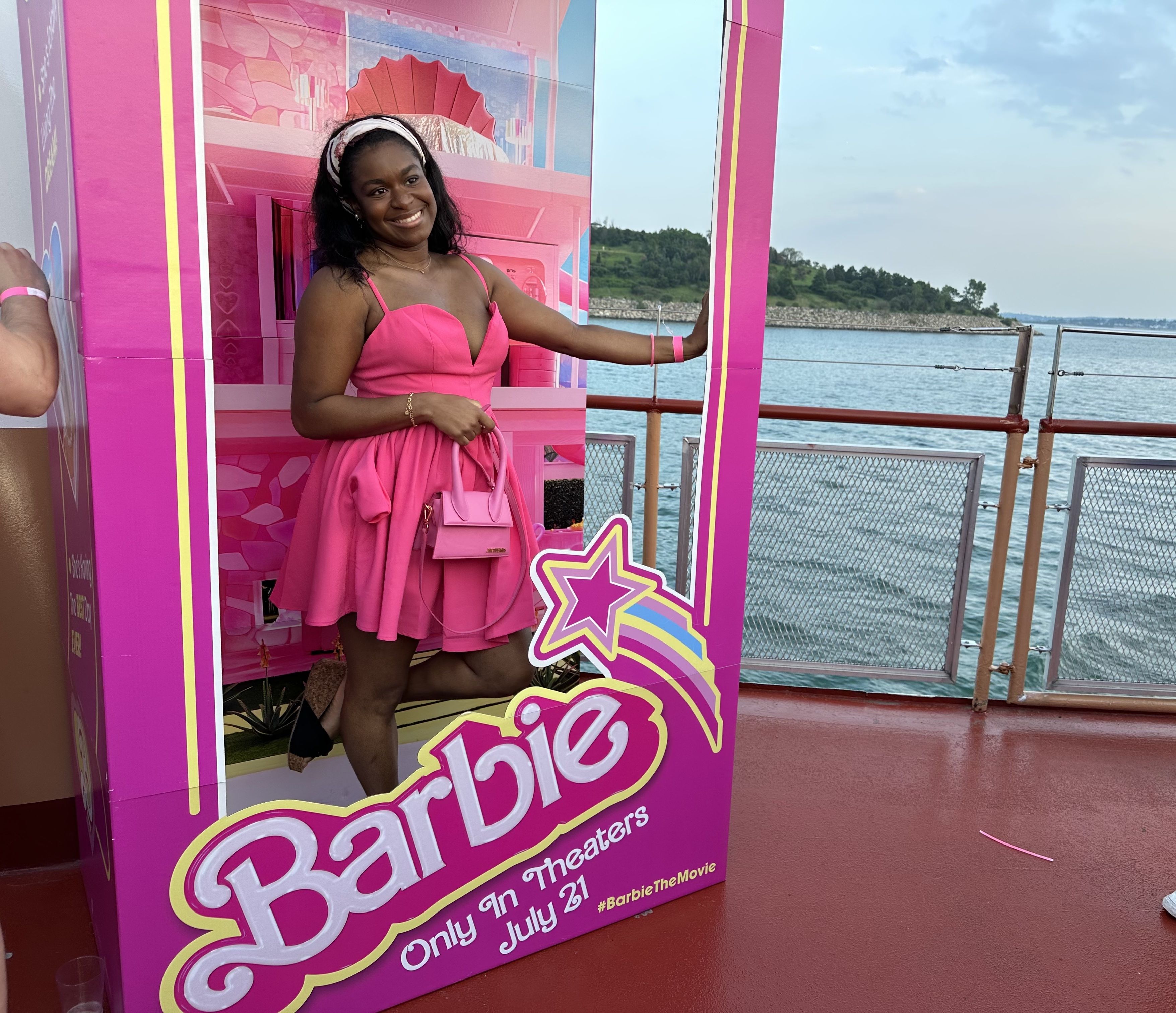 Jasmine Bacchus, a Black woman, poses for a photo inside the life-size Barbie box on the Barbie boat cruise. Jasmine wears a pink dress and small pink purse.