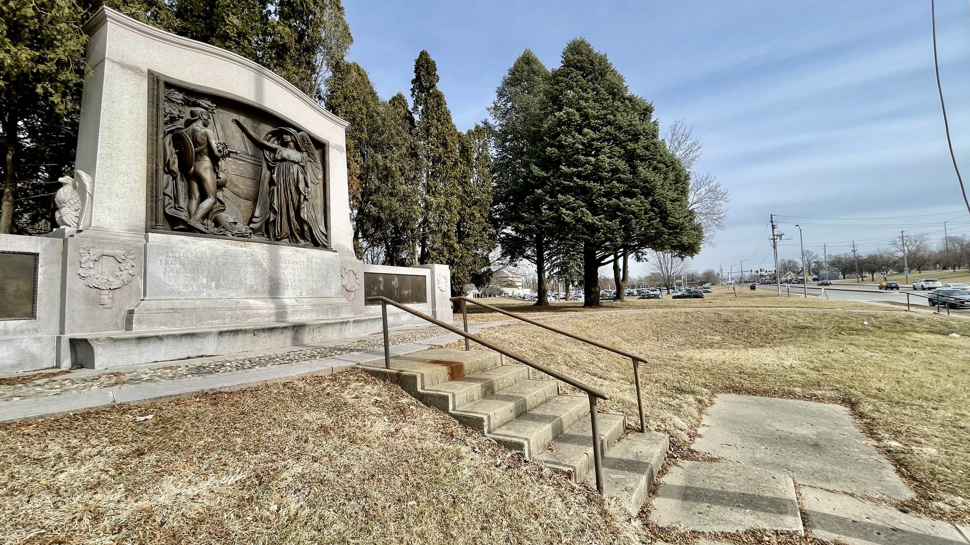 Wide view of a stone monument with a bronze relief sculpture depicting two figures, set beside steps with a metal handrail, surrounded by trees and a road with cars in the background under a blue sky.