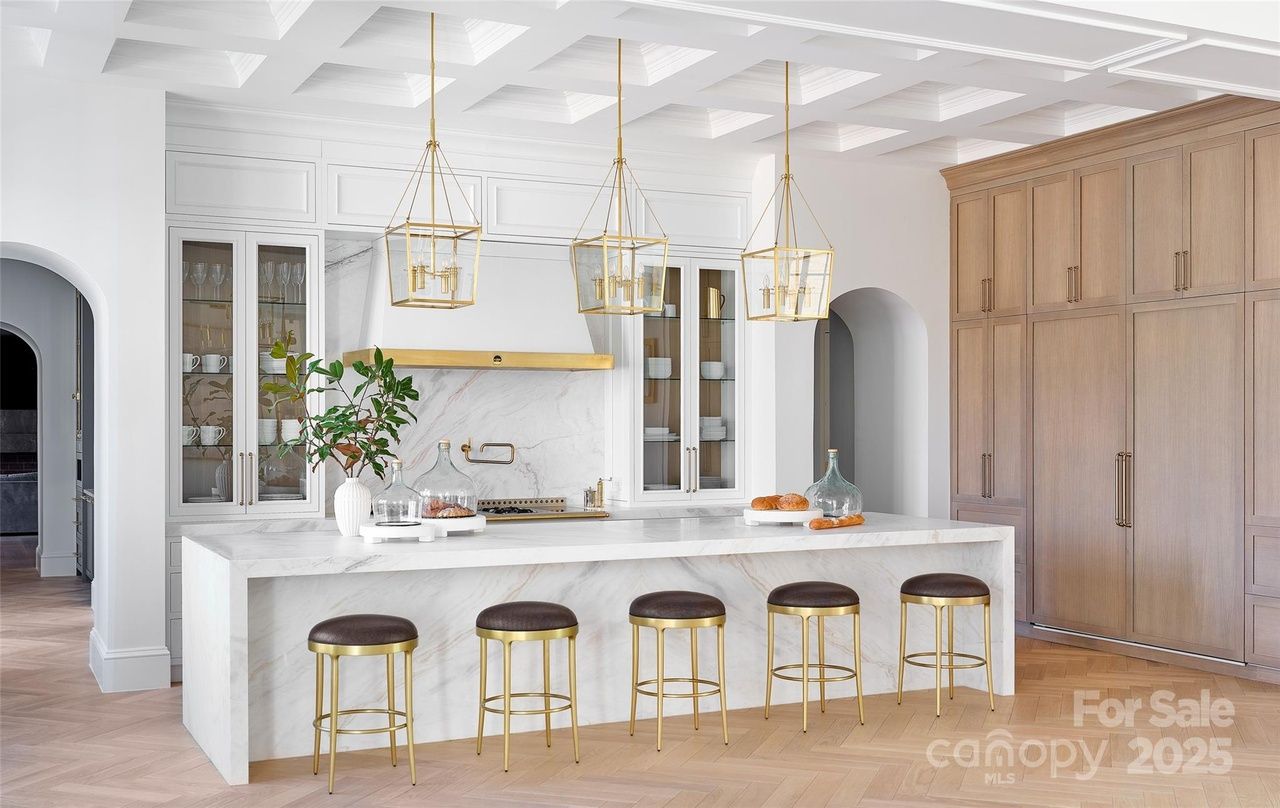 Modern kitchen with white marble island, five gold and brown stools, three gold pendant lights, white cabinetry with glass doors, wooden wall cabinets, and light parquet flooring.