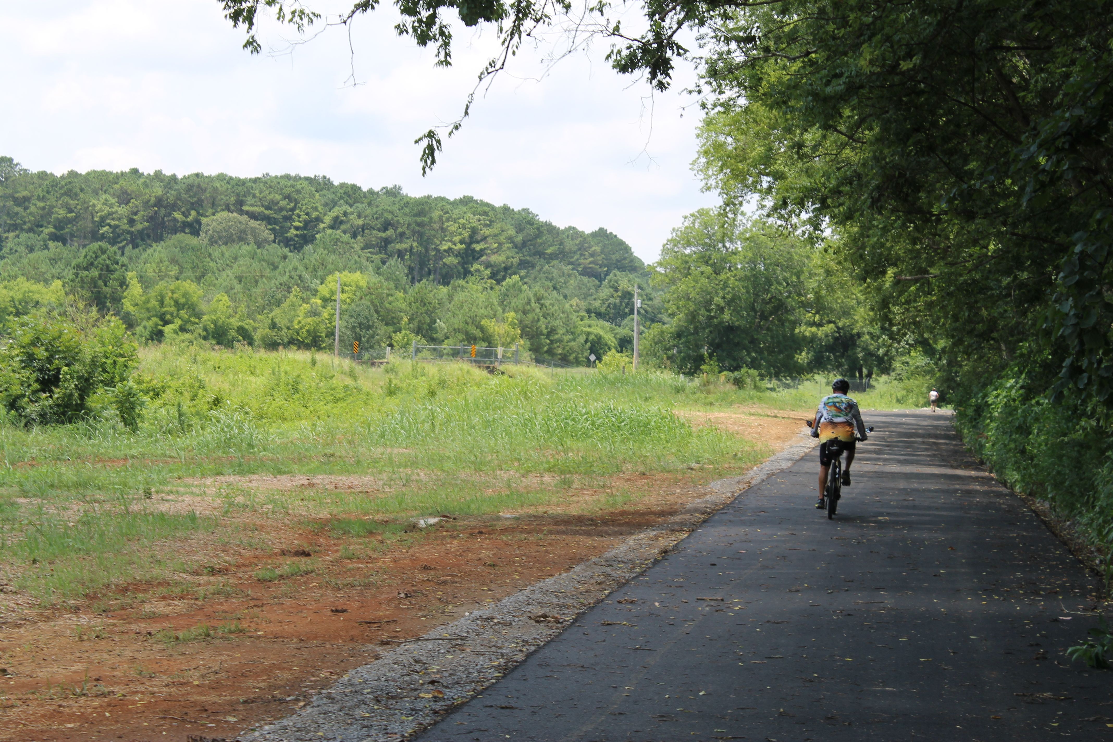 Paved trail winds through a grassy, wooded landscape with distant hills. A cyclist in a colorful shirt rides away on the right; another rider appears farther down, framed by overhanging branches.