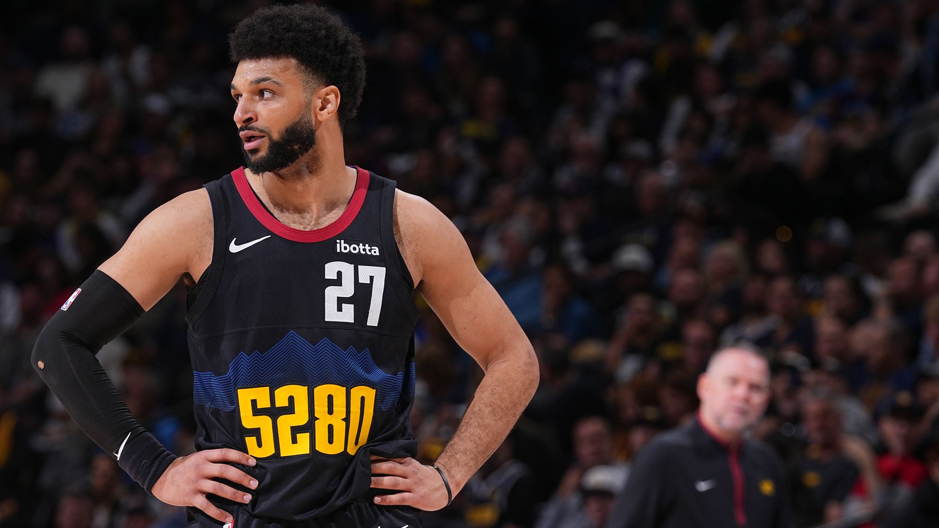 A basketball player in a black jersey with the numbers 27 and 5280 on the front of the jerseys stands with his hands on his waist. Behind him, a massive crowd is visible but blurry. 