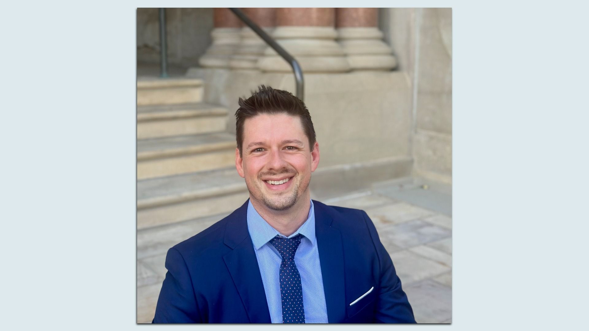 Smiling man with short dark hair wearing a blue suit, light blue shirt, and navy tie with small dots, sitting outdoors near stone steps and beige columns.