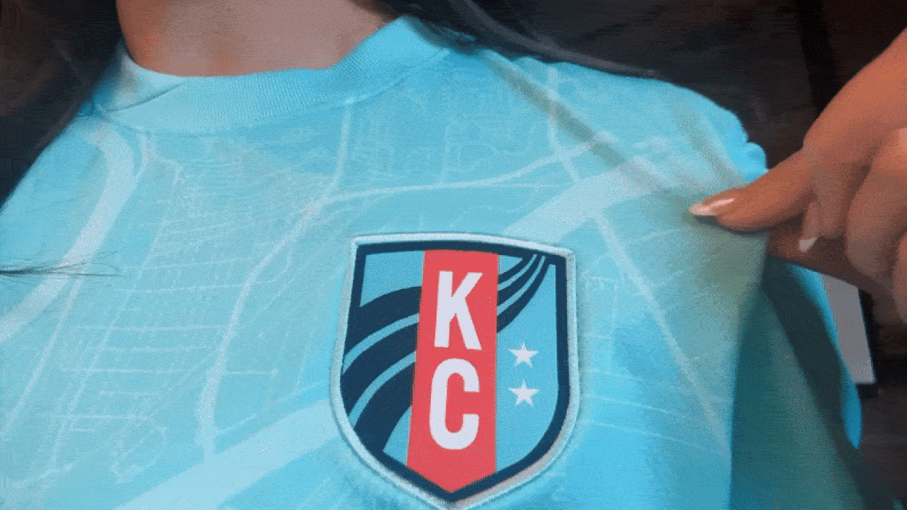 Light blue shirt with a shield-shaped KC logo featuring blue, red, and white colors and two stars; map pattern printed on the fabric.