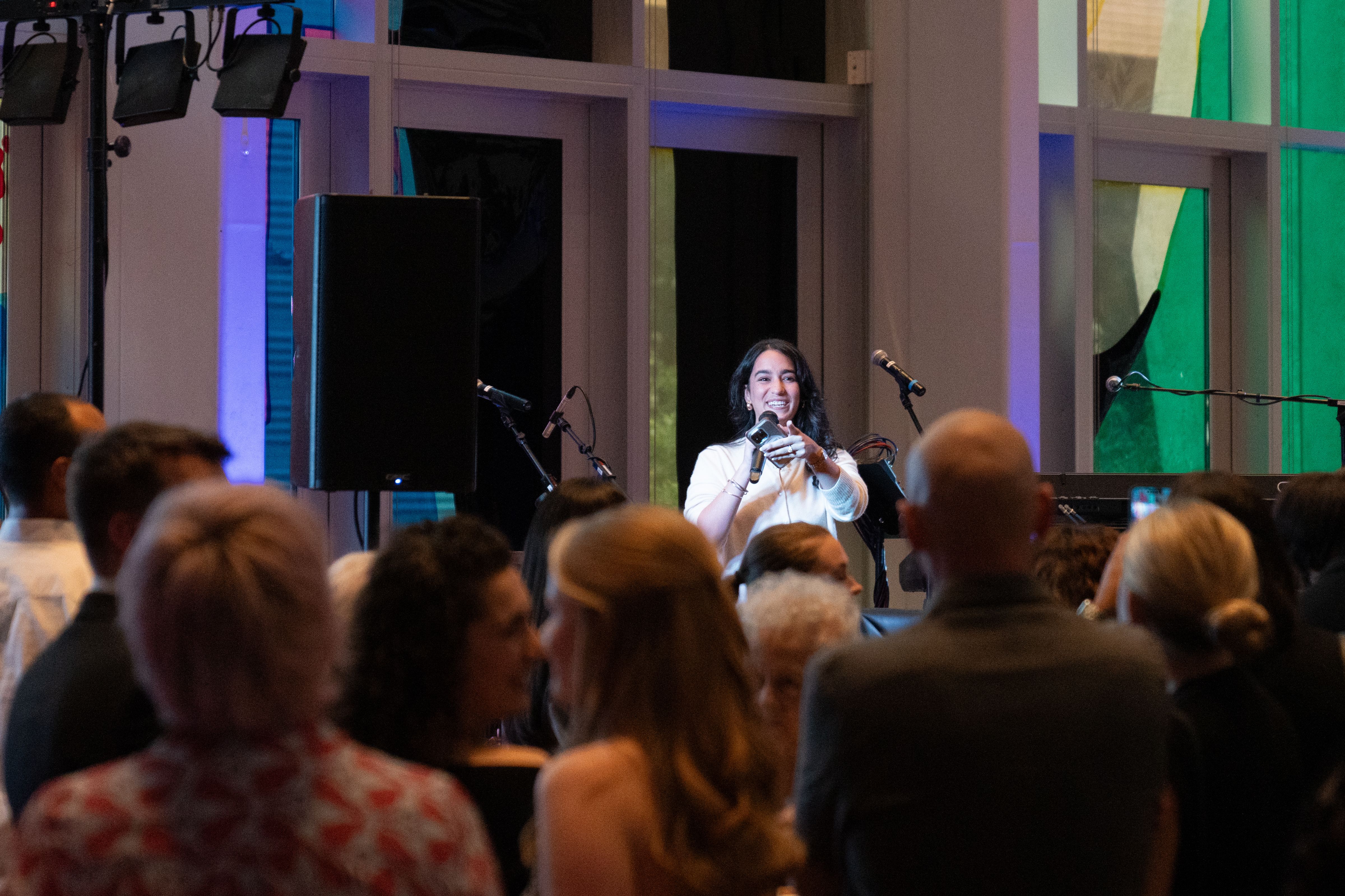 A woman in white speaking into a microphone and smiling at an indoor event, with a crowd of people watching her. Colorful stained glass windows and speakers are visible in the background.
