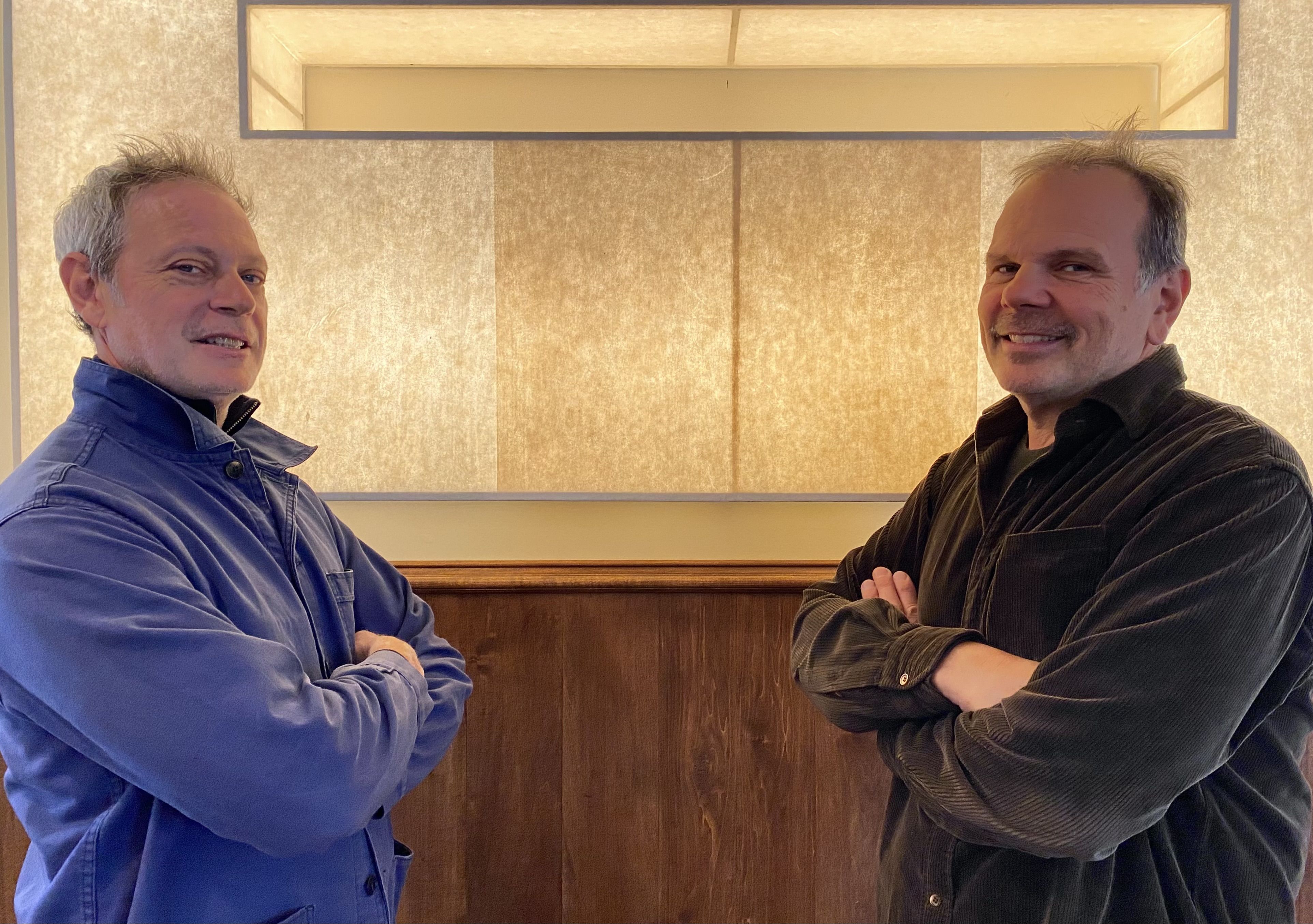 Two middle-aged men facing each other with arms crossed and smiling, one wearing a blue jacket, the other a dark brown shirt, standing indoors against a beige wall with wood paneling.