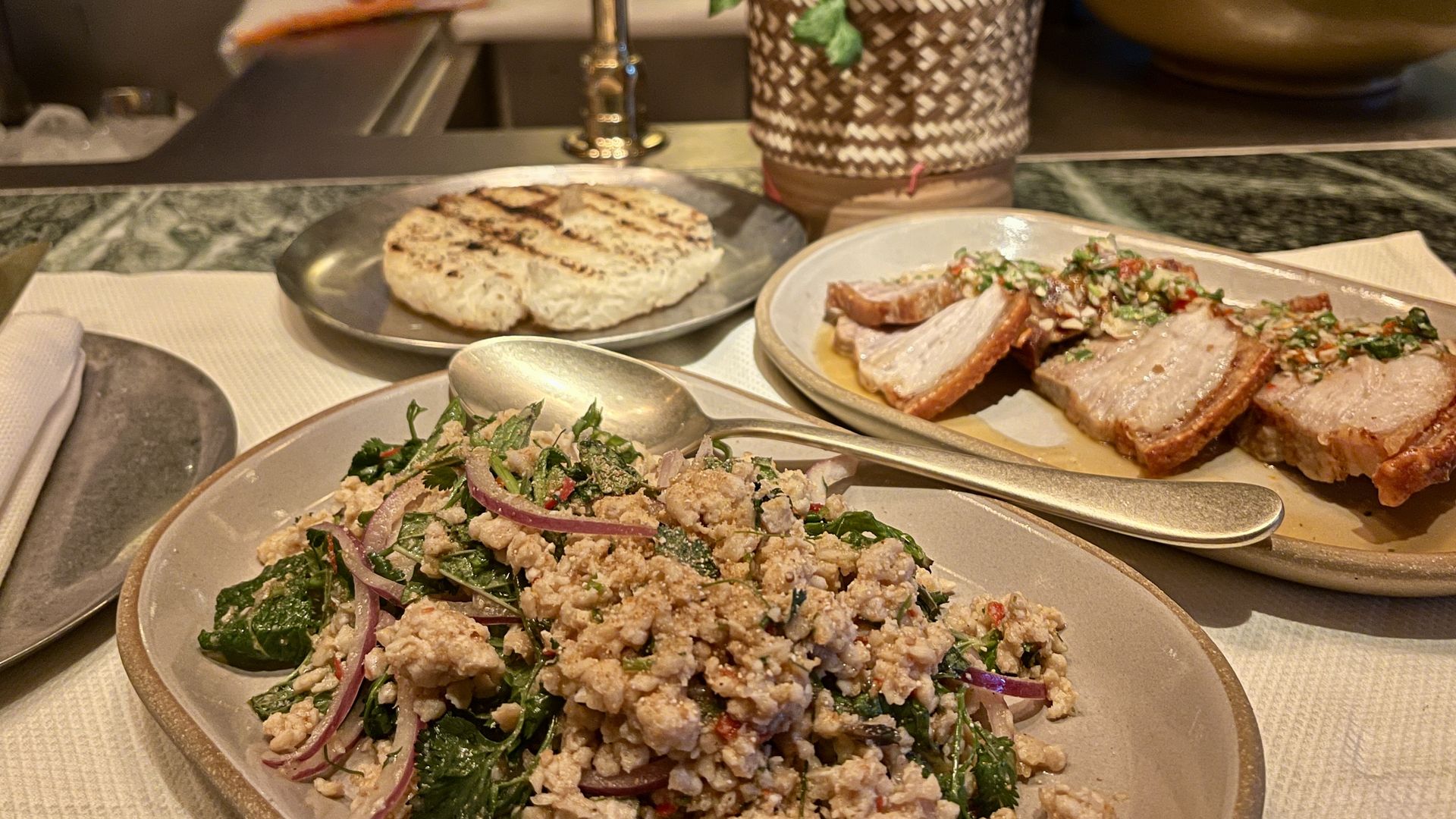 Close-up of a meal at a restaurant, featuring minced meat salad with herbs and red onions, crispy pork slices topped with sauce on a plate, and grilled sticky rice on a metal plate, with a plant in a woven pot.