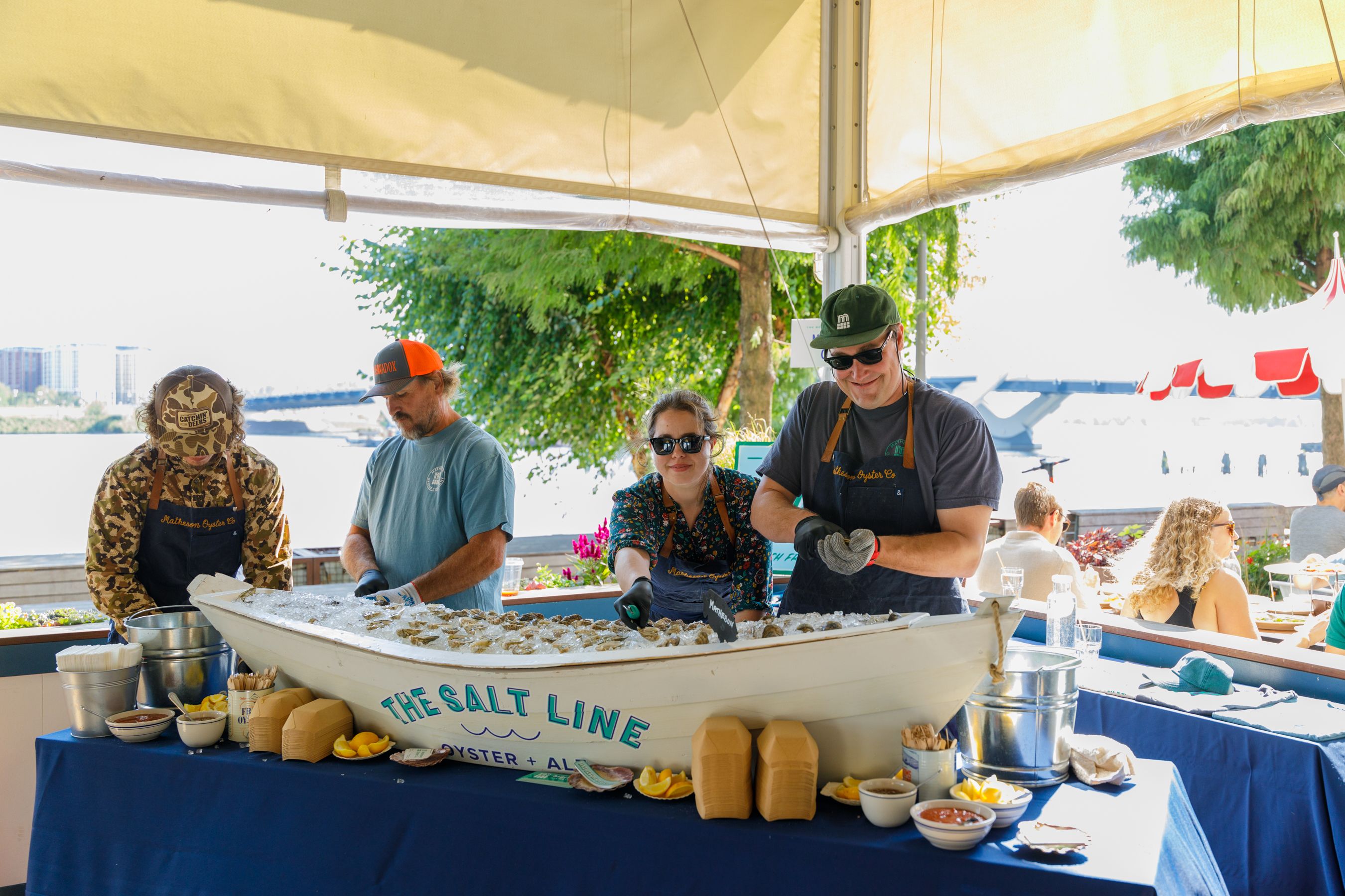 Under a yellow canopy, workers shuck oysters at a sunny outdoor stand. Four people in aprons and caps work over ice, with bowls, lemons, and sauces; trees and water in the background.