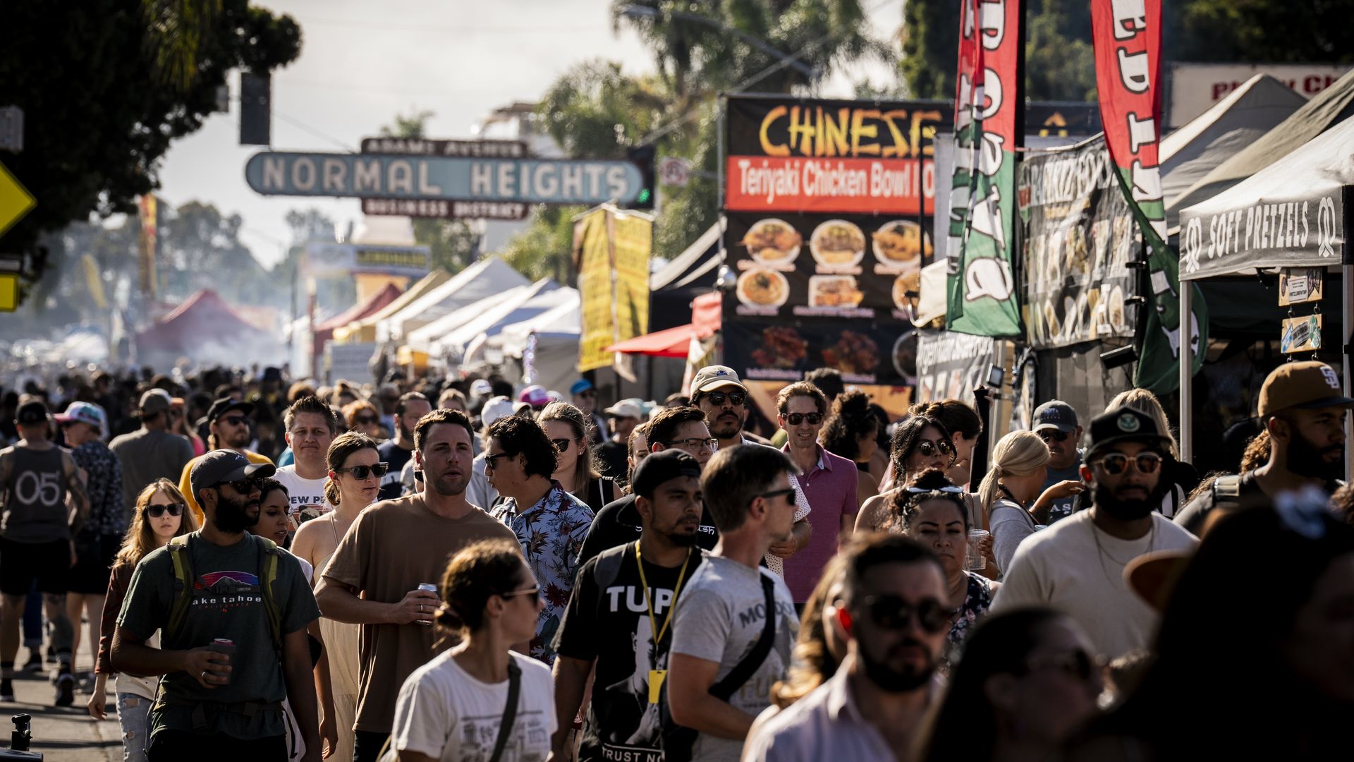 Crowded street fair in Normal Heights Business District with people walking past food stalls on a sunny day.