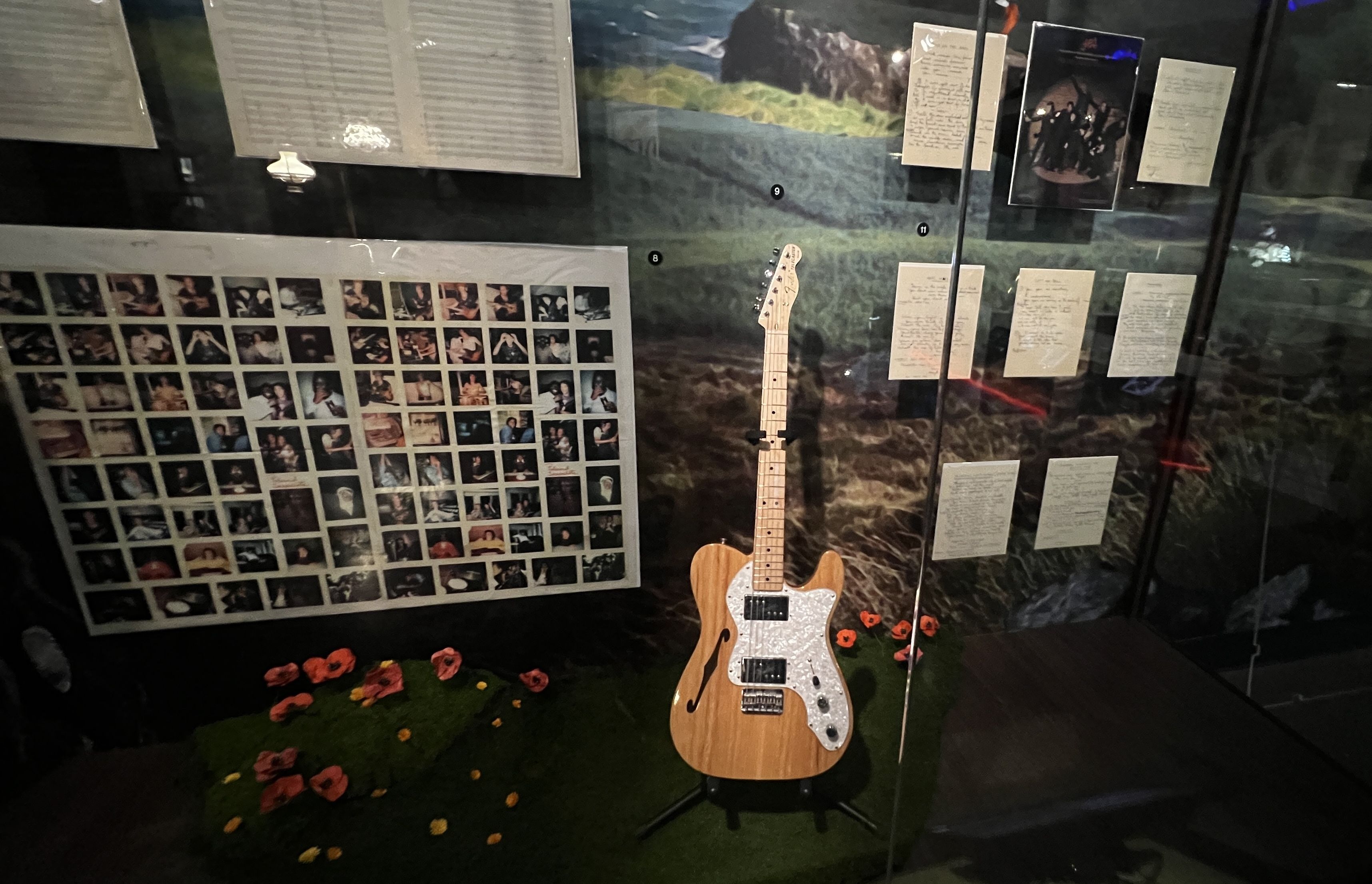 Exhibit case showing a light wood electric guitar on a stand, a left wall collage of small photos, handwritten notes on glass panels, and red and yellow flowers on the floor.