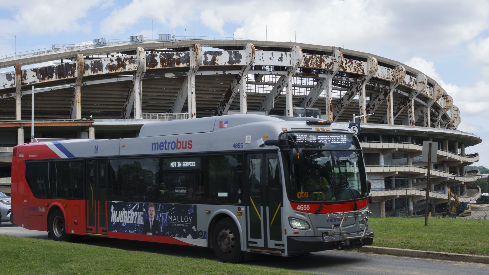 Gray and red Metrobus, not in service, parked near an old, rusted, RFK Stadium under a cloudy sky.