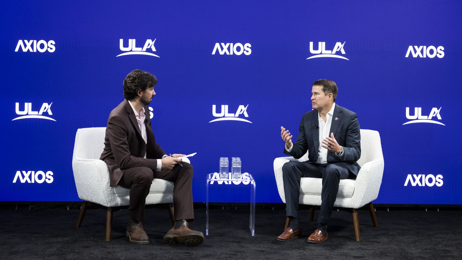 Two men in suits sit on white armchairs on a black carpeted stage with a blue background displaying "ULA" and "AXIOS" logos. A small clear table with two glasses of water is between them.