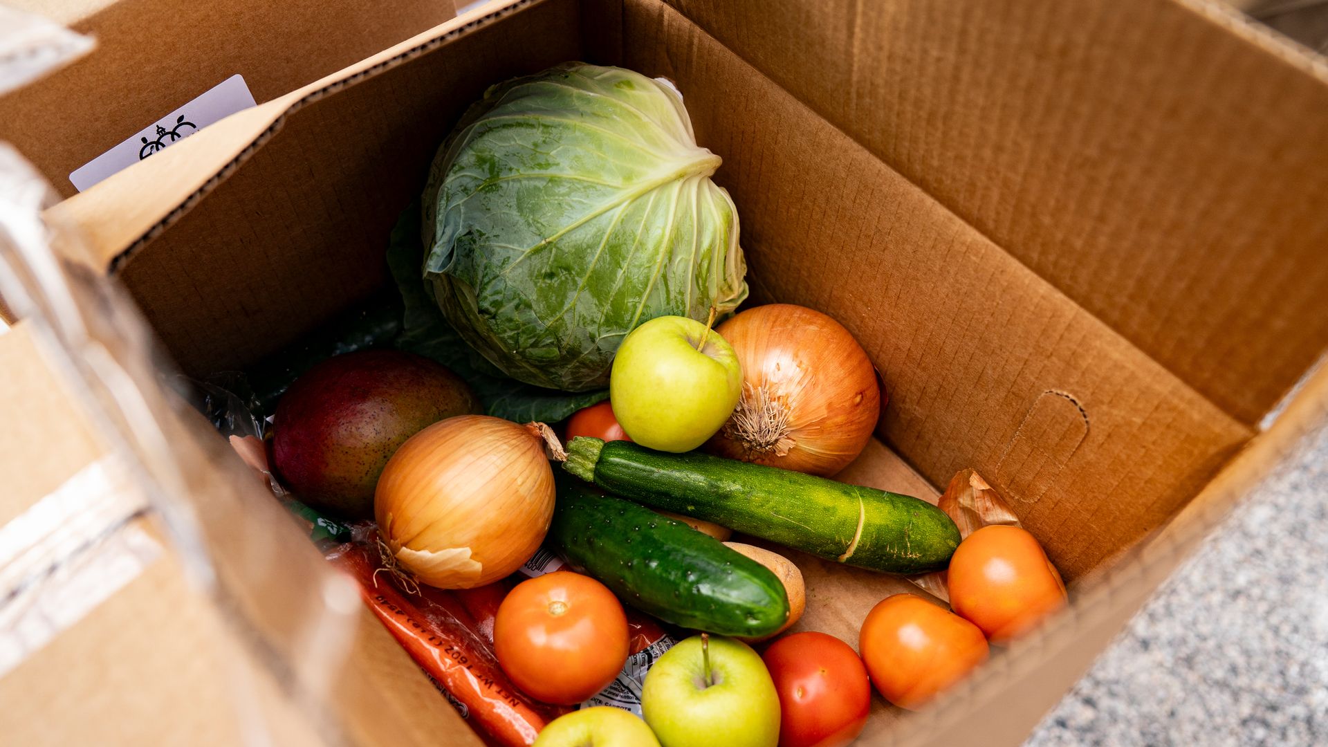A cardboard box holds several produce items, including a head of lettuce, onions, apples and cucumbers. 
