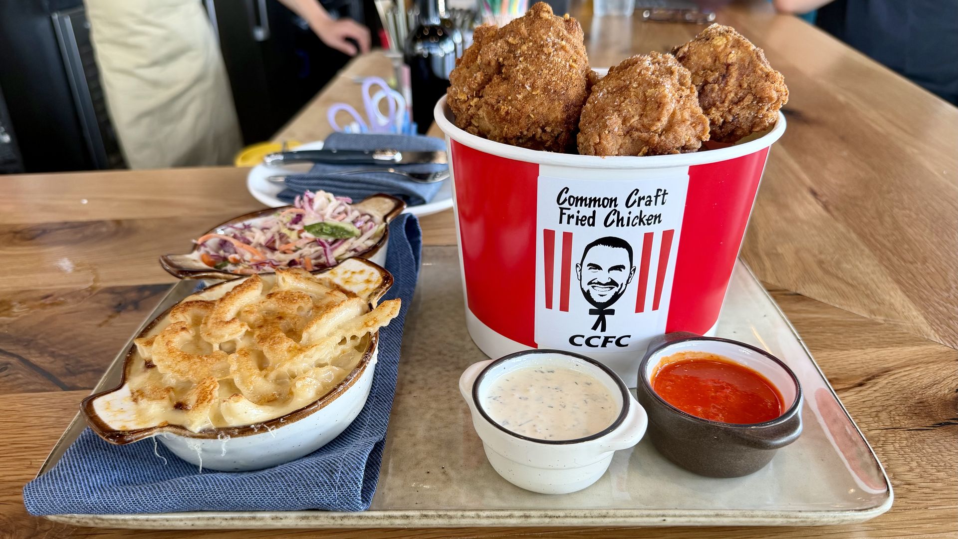 Red and white bucket of fried chicken labeled Common Craft Fried Chicken sits on a wooden table with macaroni and cheese, coleslaw, and three dipping sauces; kitchen in the background.