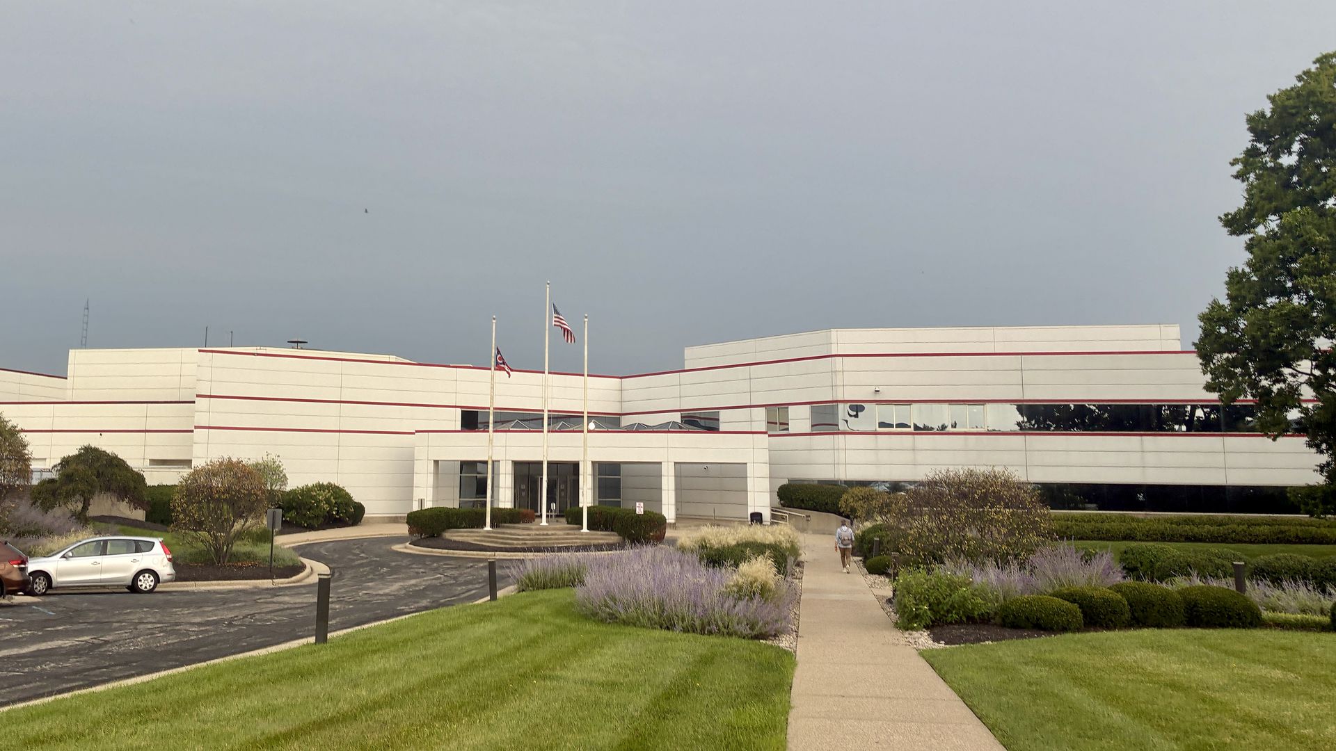 White modern office building with red trim under a pale blue sky. Flagpoles bear flags in front of the glass entrance. A lavender-lined sidewalk leads to the doors, with cars on the left and trees on the right.