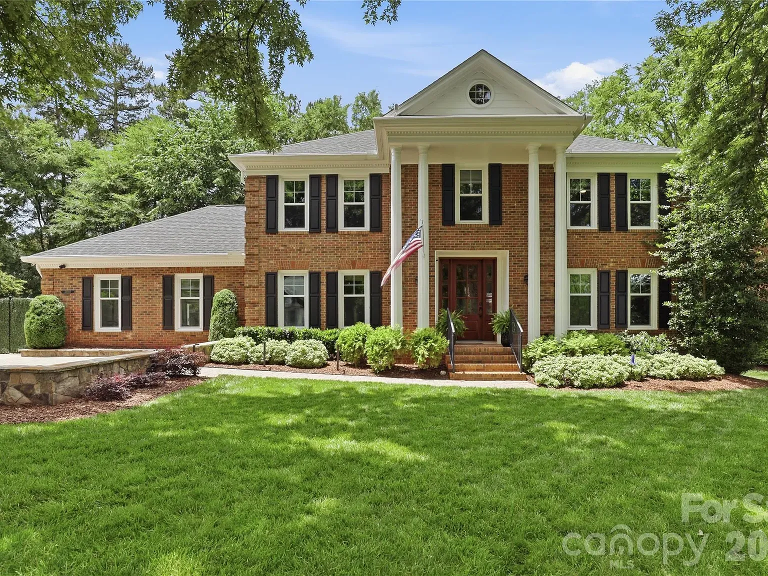 Front view of a two-story red brick house with white columns, black shutters, and a gabled pediment. A flag on a pole stands near the maroon front door, with a manicured lawn and shrubs.