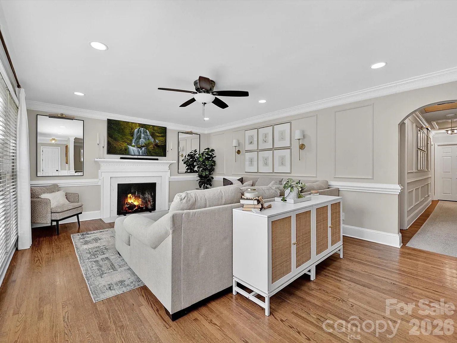 Bright living room with light beige walls, hardwood floors, gray sofa, white fireplace with fire, two large mirrors, TV showing waterfall, and wooden cabinet with plants and books.