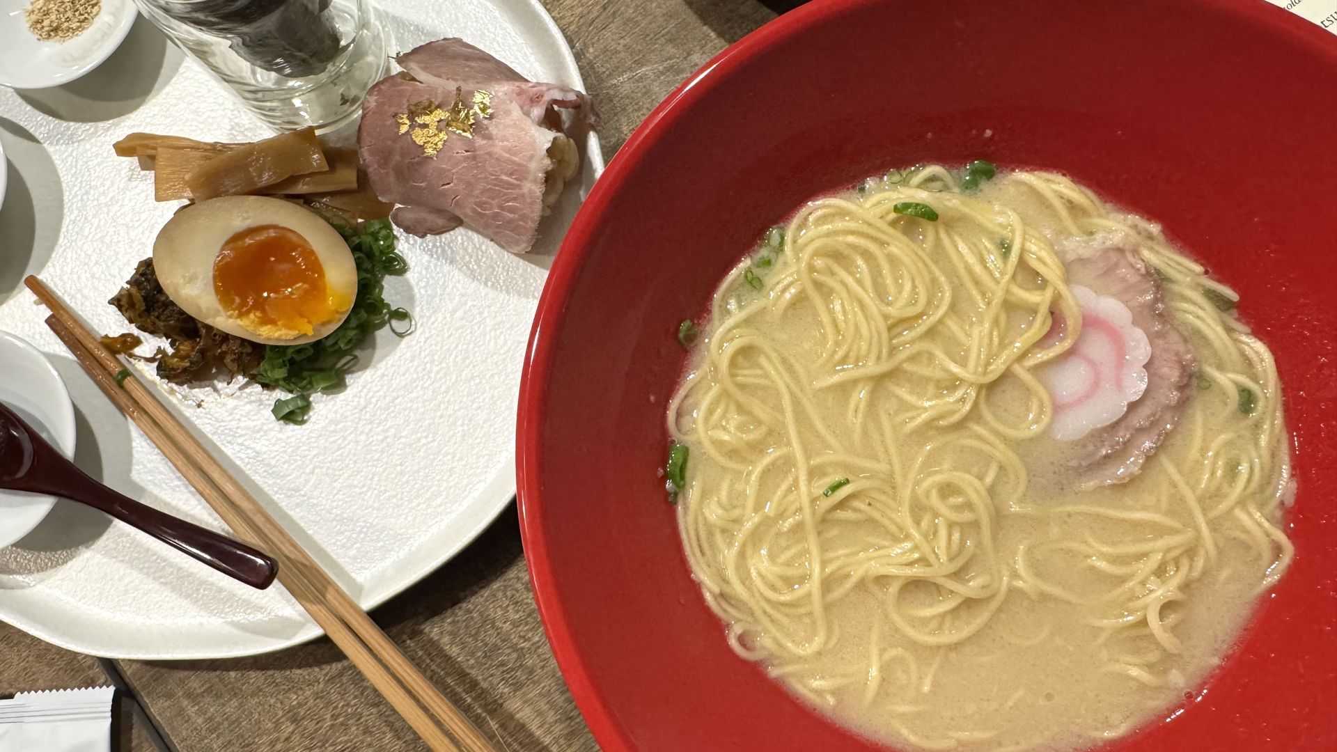 Red bowl of ramen with noodles, broth, sliced pork, green onions, and a pink-and-white narutomaki fish cake, beside white plate with half a soft-boiled egg, bamboo shoots, chopped green onions, and sliced meat topped with gold flakes.