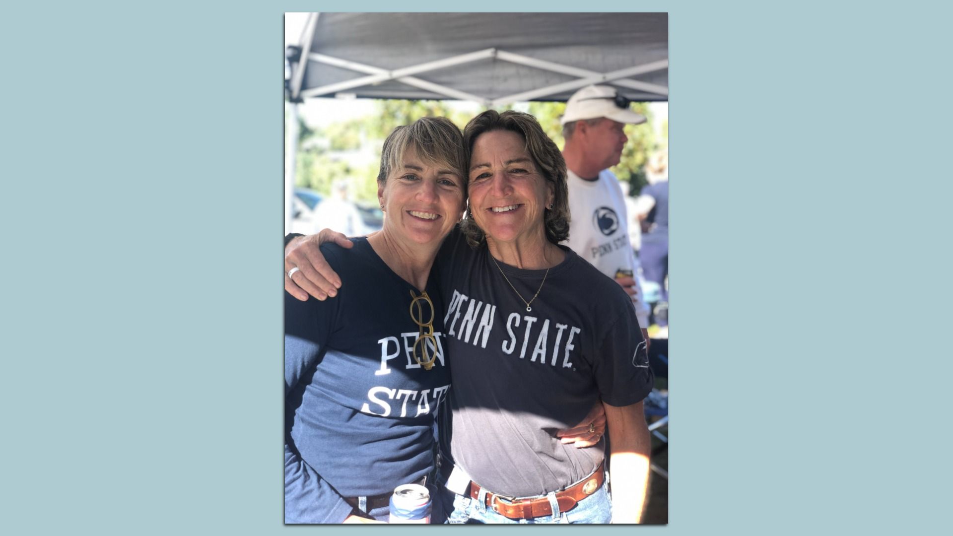 Two smiling women wearing Penn State shirts pose with their arms around each other under a canopy at an outdoor event; a man in a white cap stands behind them.