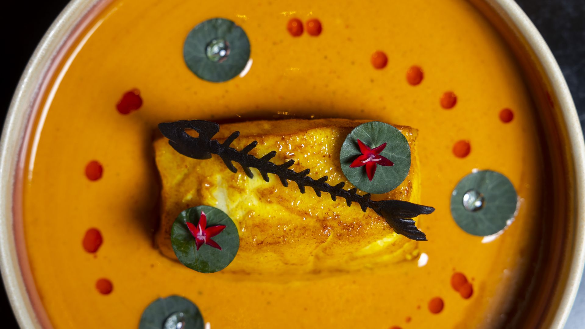 A plated dish with a piece of golden-brown fish in bright orange sauce, decorated with green leaves, small red flowers, and a black fishbone-shaped garnish on top.