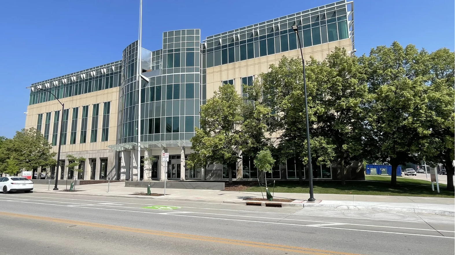 Modern beige and green glass building with large windows, trees along the sidewalk, and clear blue sky above on a sunny day.