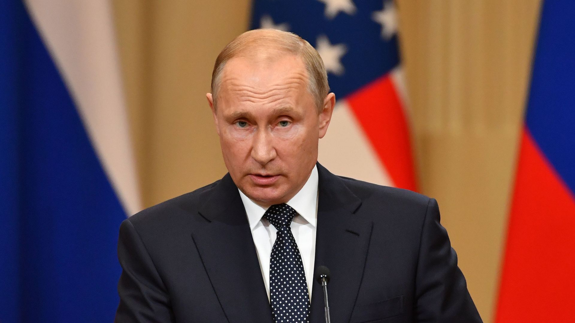 Vladimir Putin looks caught off guard, standing at a podium before a U.S. flag.