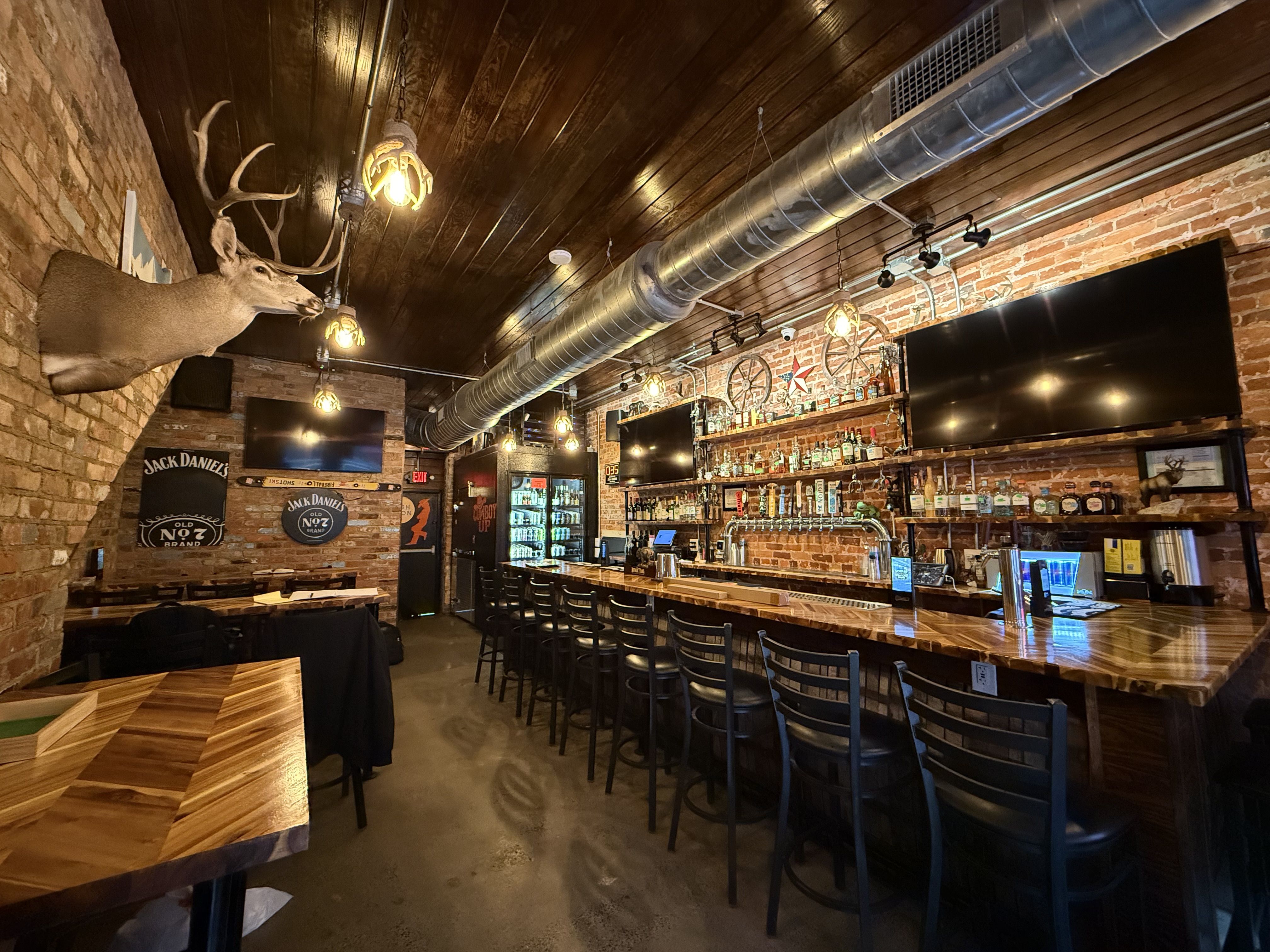 Interior of a dimly lit bar with wooden ceiling and brick walls, featuring a mounted deer head, a long wooden bar with black chairs, three large TV screens, and Jack Daniel's signs.