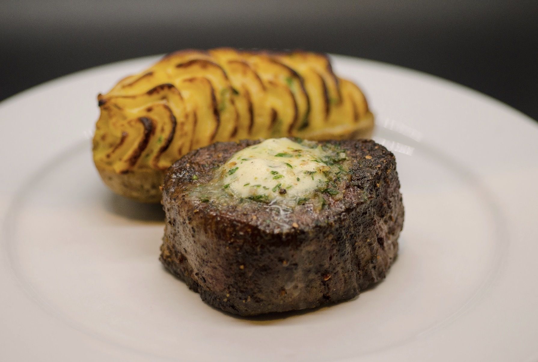 Thick seared beef tenderloin medallion topped with melted herb butter, served with a golden spiral-cut potato beside it on a white plate.