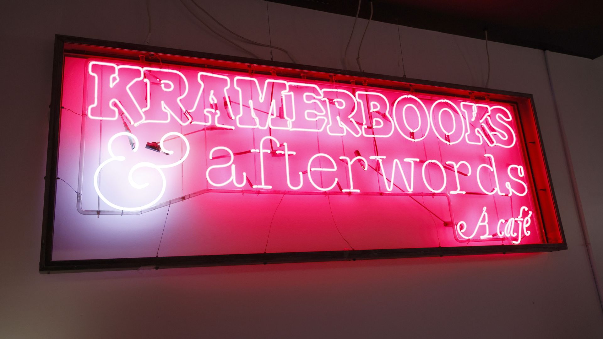 A neon pink sign framed on a dark wall, with large neon letters spelling a phrase ending in BOOKS and smaller cursive text reading Cafe in the bottom right.