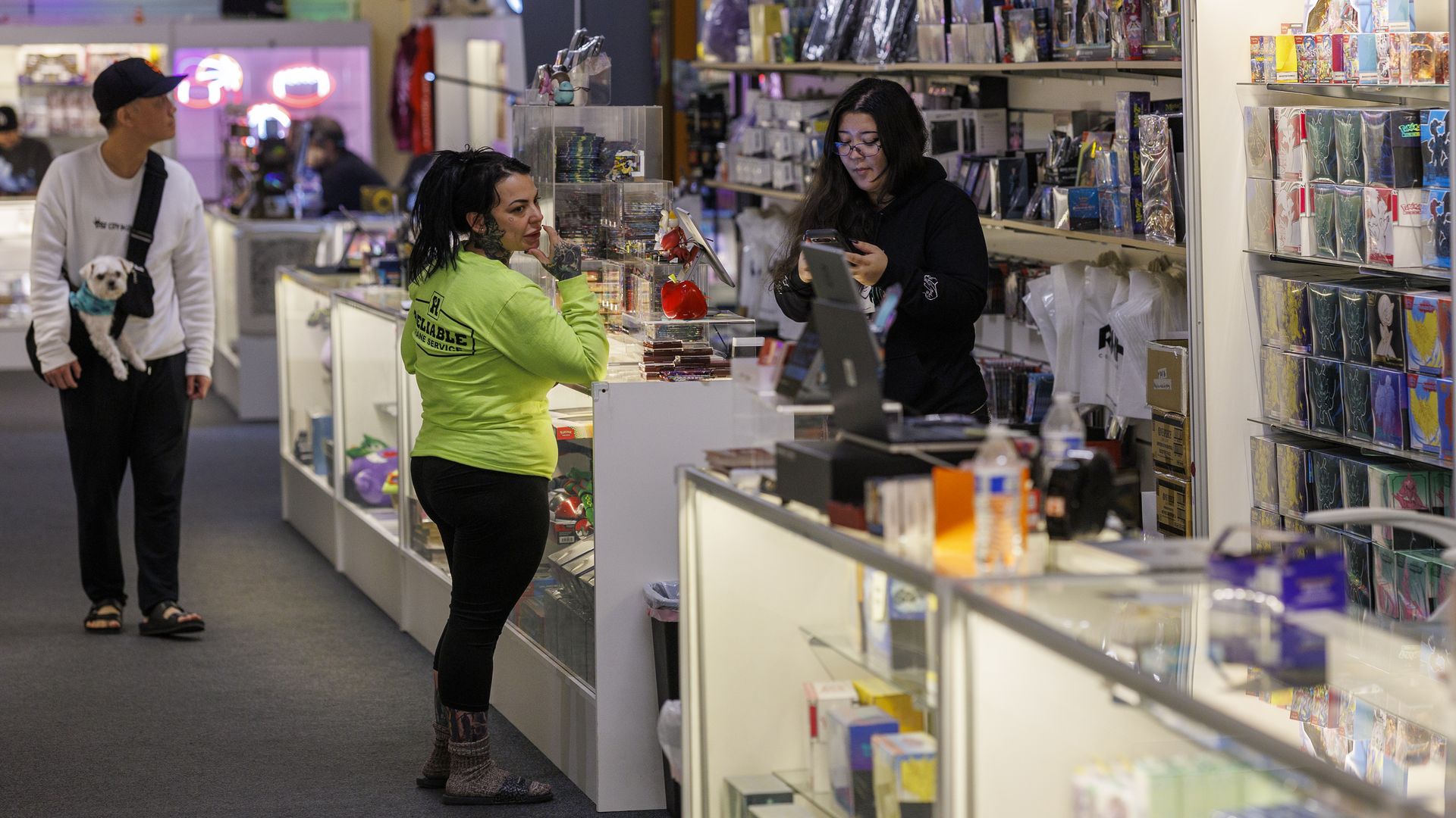 Inside a colorful shop with glass display cases; a tattooed person in neon green talks to a clerk at the counter as a person with a small white dog passes by; wall shelves hold cards and prints.