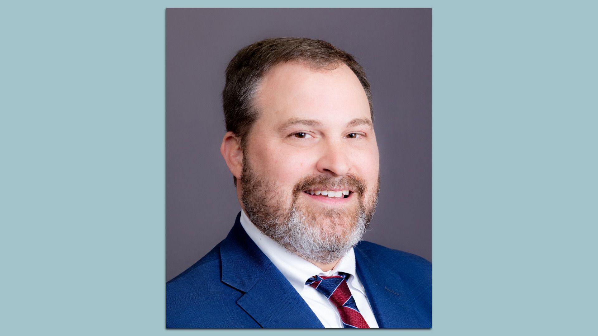 A headshot of a smiling individual with a beard wearing a dark blue suit and red and blue striped tie.