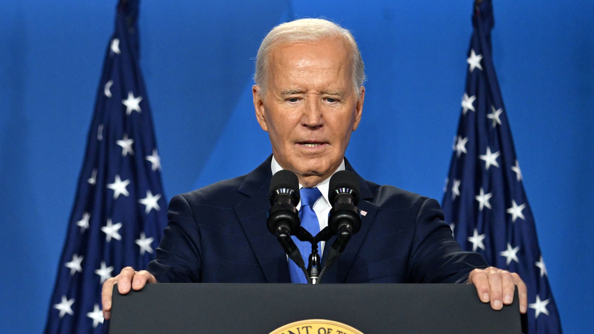 President Biden, wearing a blue suit and gripping a podium in front of American flags and a blue wall.