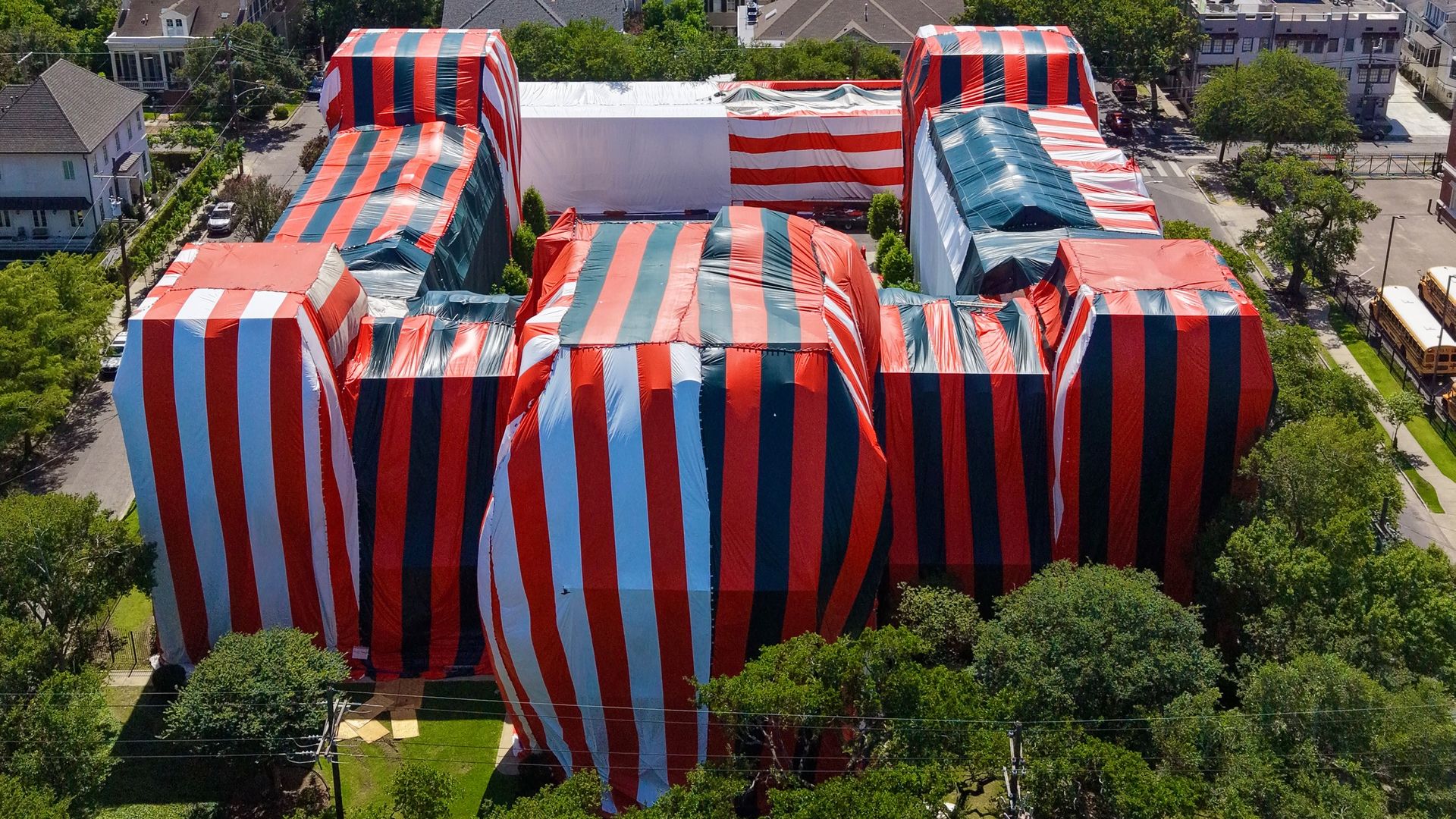 An aerial view of a suburban city with green trees and houses; in the foreground, a festival site features large red, white, and blue striped tents arranged in a maze-like layout.