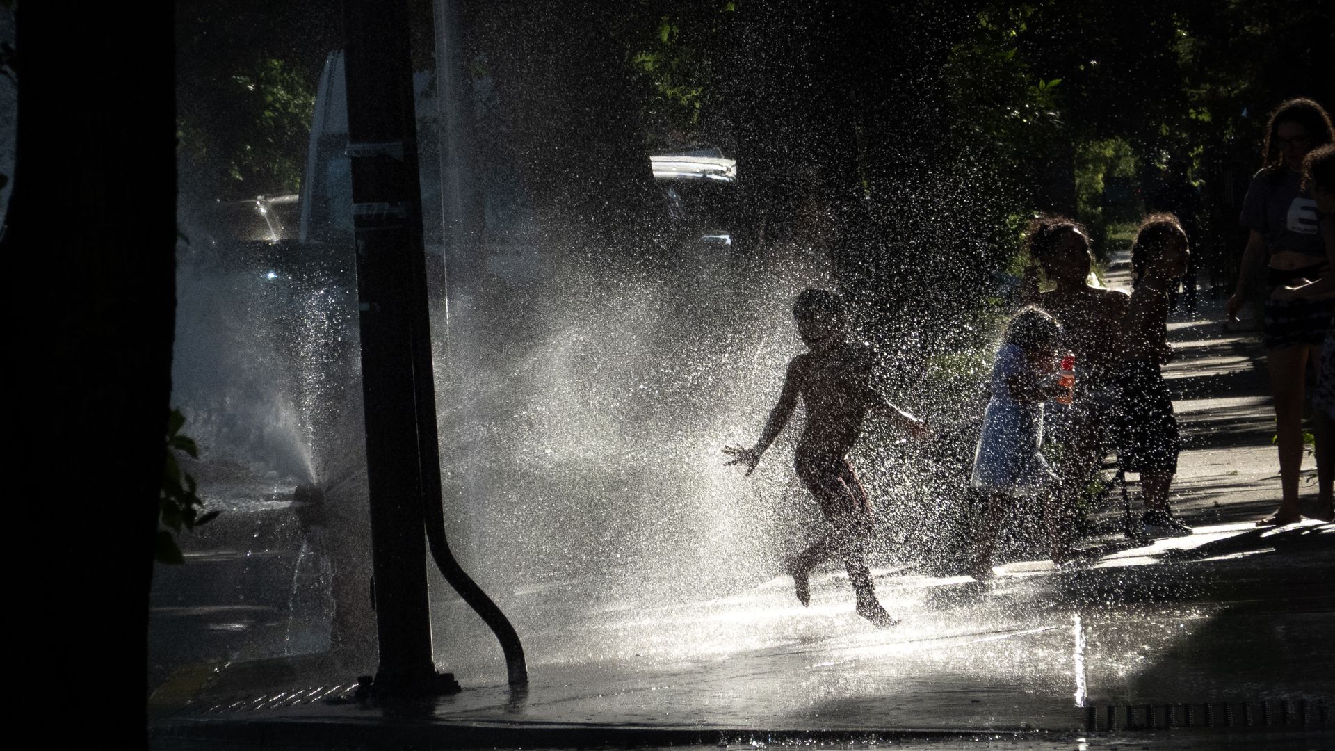 Silhouetted people walk through a heavy spray from a fountain on a sunny street; sparkling water droplets fill the air as trees and dark structures loom in the background.