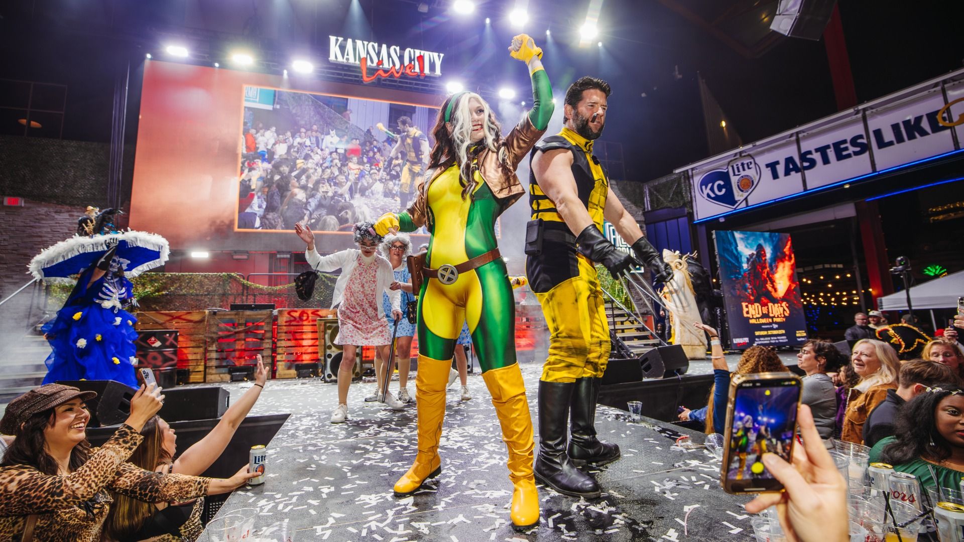 Cosplayers pose center stage at a lively indoor event, white-haired woman in green-yellow against a yellow-black man. Confetti on the floor; crowd cheers; "Kansas City Live" sign overhead.
