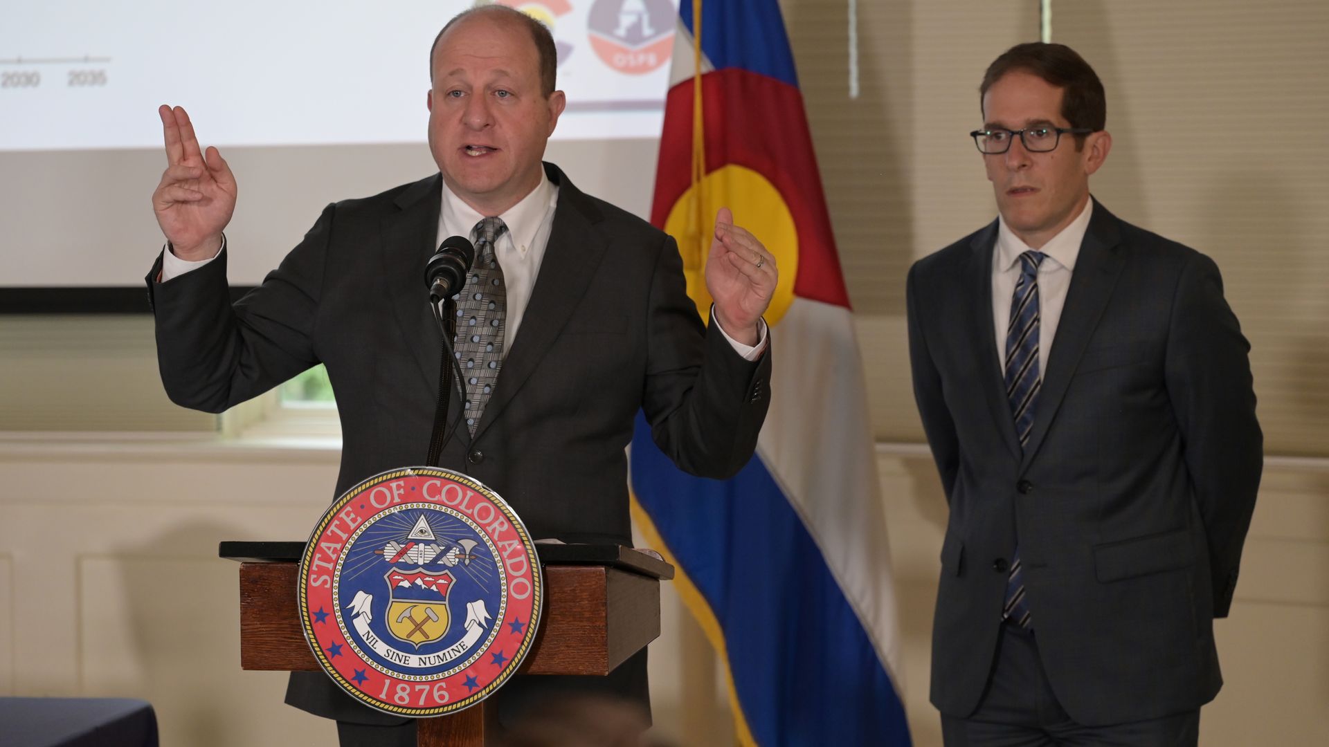 Gov. Jared Polis speaks with hands raised at a podium.