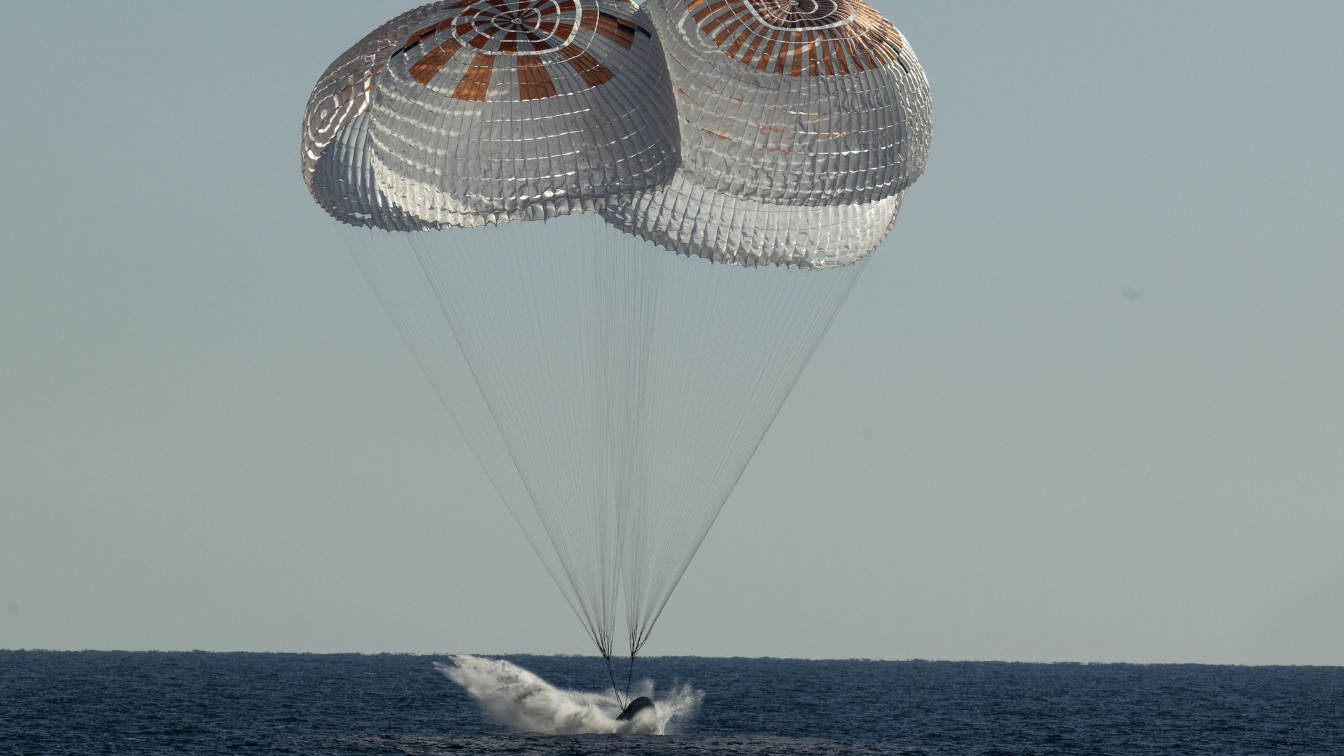 A capsule splashes down in the ocean, sending up white spray under orange and white parachutes as the Sun is starting to set