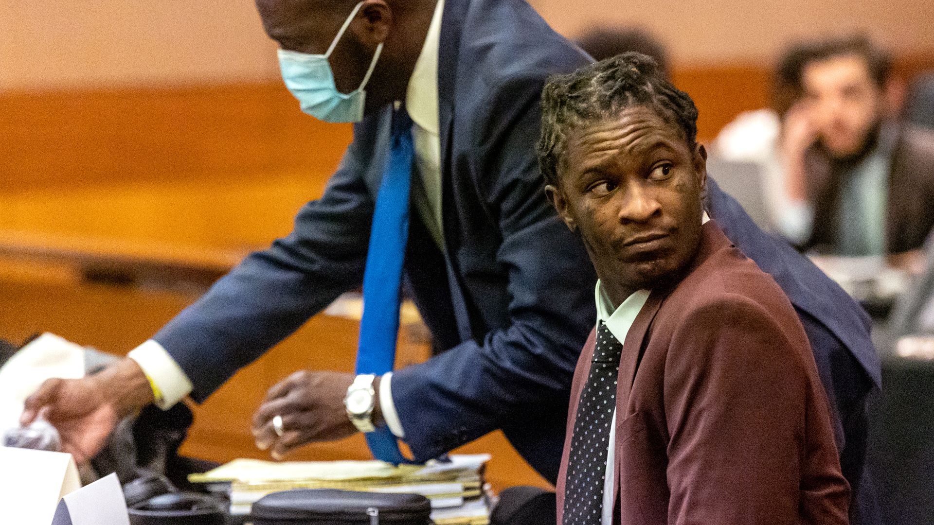 Young Thug sits at a defendant's desk in a courtroom and looks over his shoulder as his attorney wearing a surgical mask picks up a paper