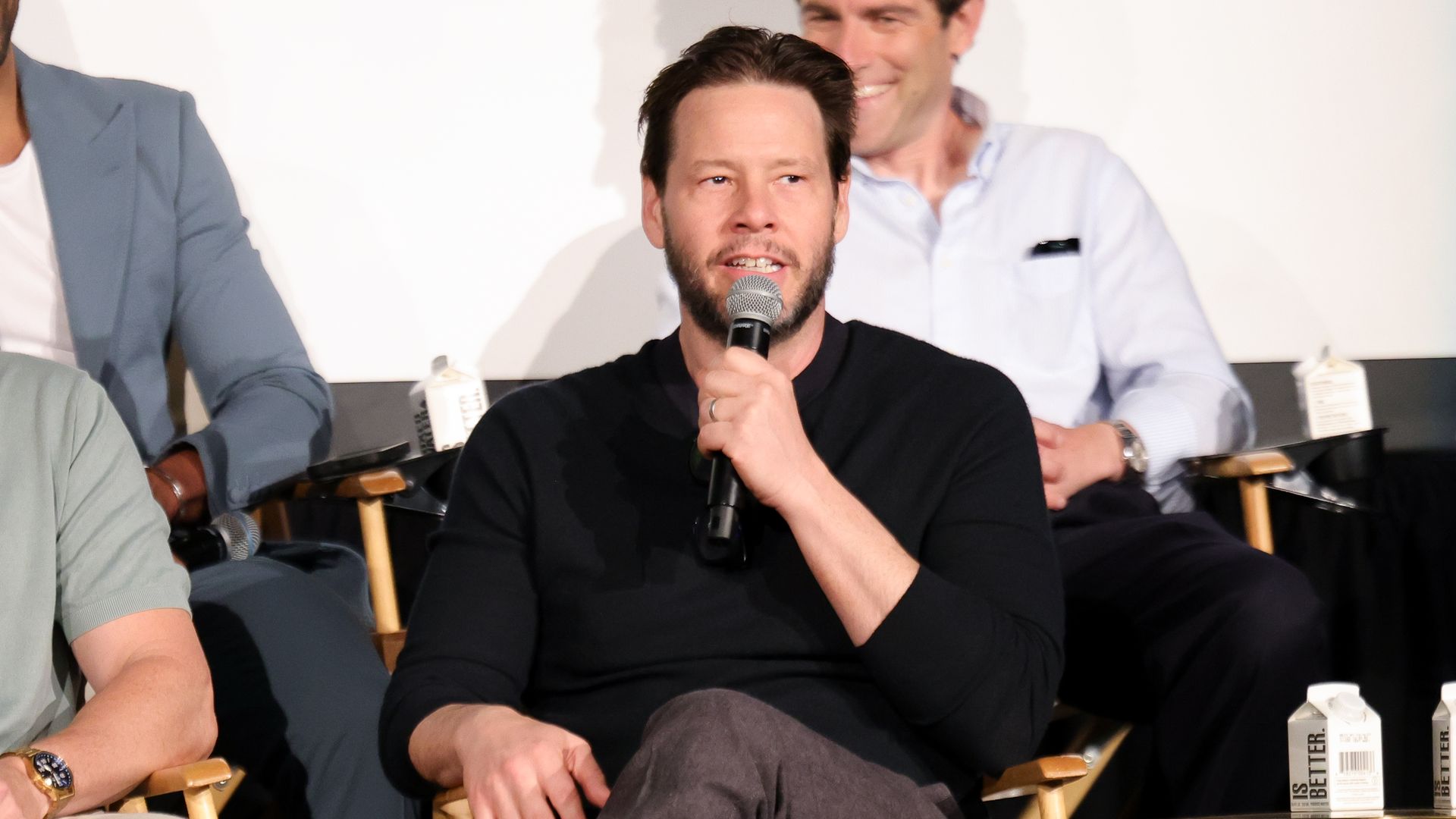 A man with brown hair and beard wearing a black sweater and gray pants speaks into a microphone while seated. Another man in a light blue shirt sits smiling behind him on a stage panel.